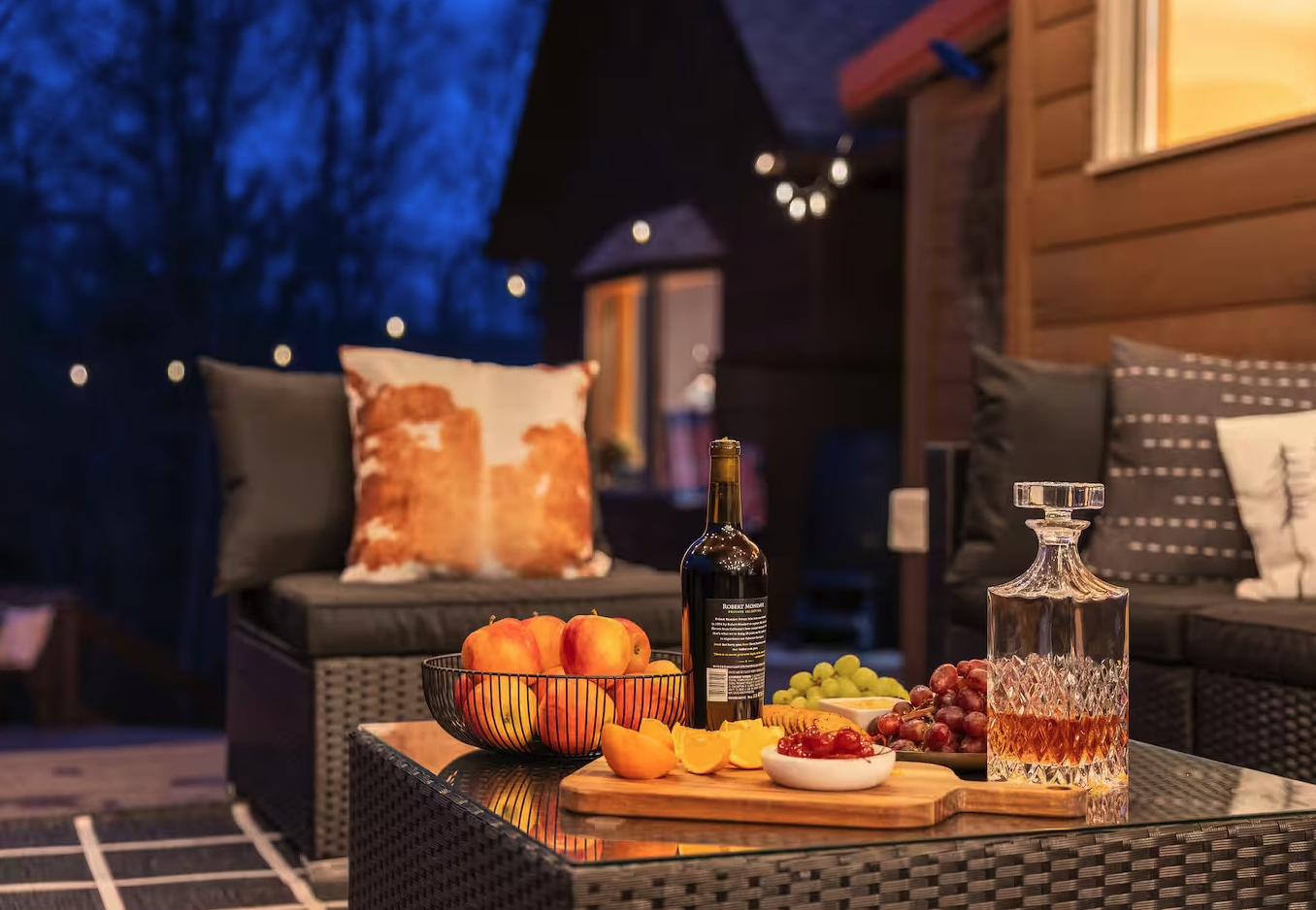 Nighttime outdoor patio with a wicker table holding a bottle of wine, a decanter, grapes, sliced peaches, and a bowl of jam, with cushioned seating and decorated pillows, surrounded by string lights and wooden house siding.