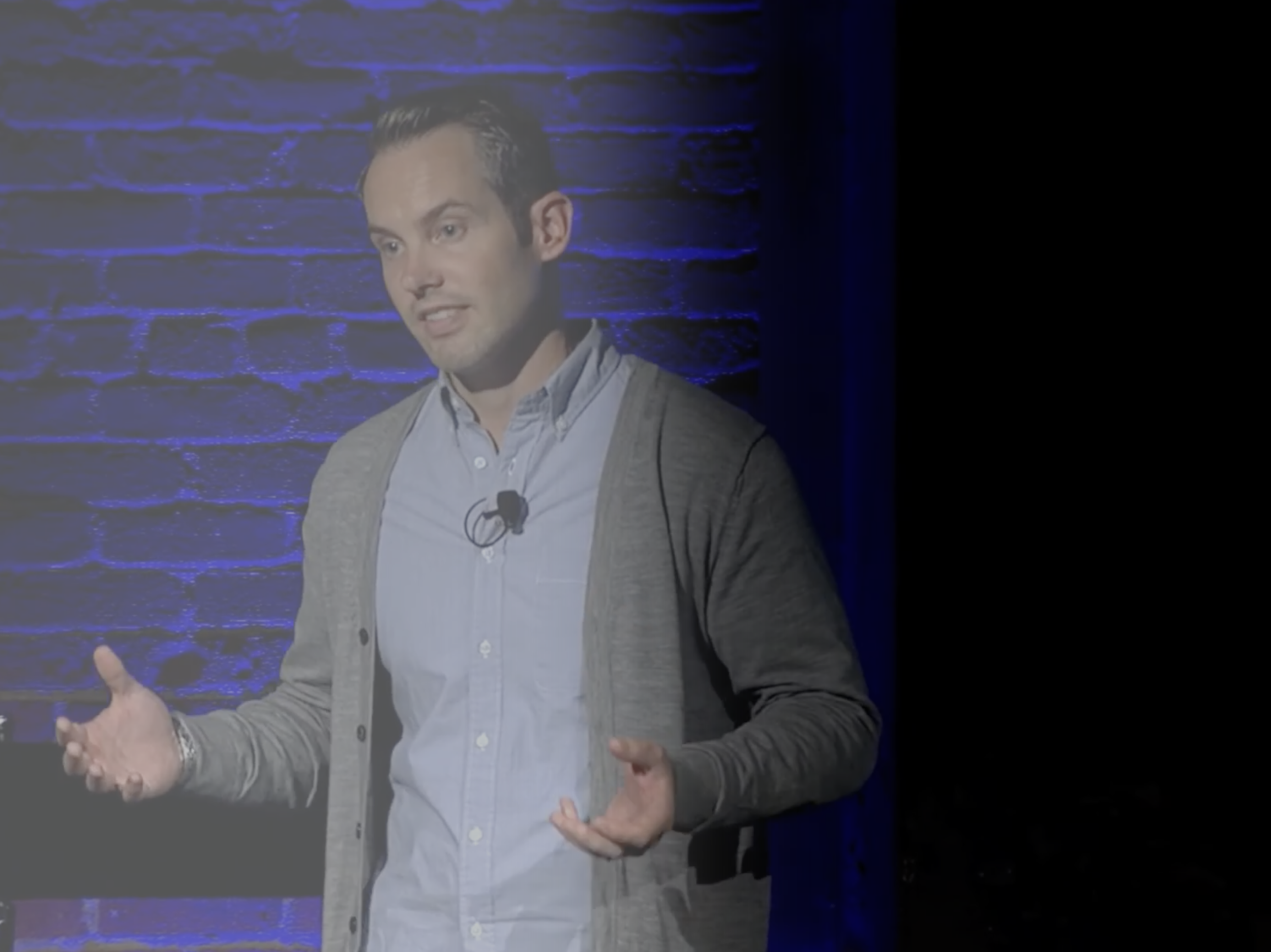 Wick Vipond giving a presentation in front of a blue-lit brick wall, wearing a button-up shirt and a gray cardigan.