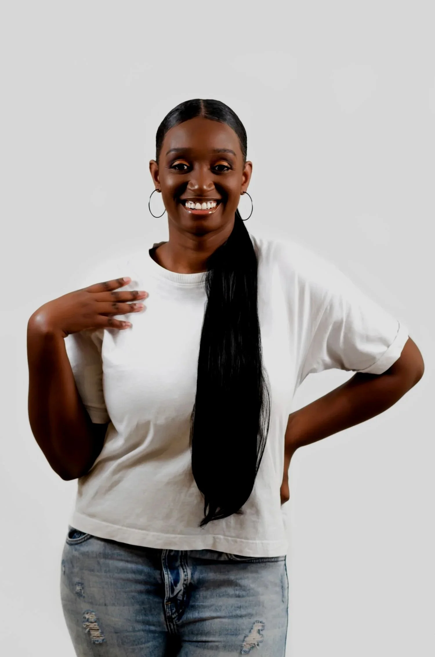Smiling woman with long black hair, hoop earrings, wearing a white t-shirt and ripped jeans, standing against a plain white background. Founder of Livequipped a Christian lifestyle blog