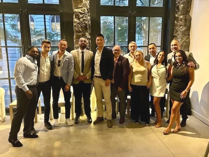 Group of fifteen people posing indoors in front of large windows, some dressed in formal or business casual attire.