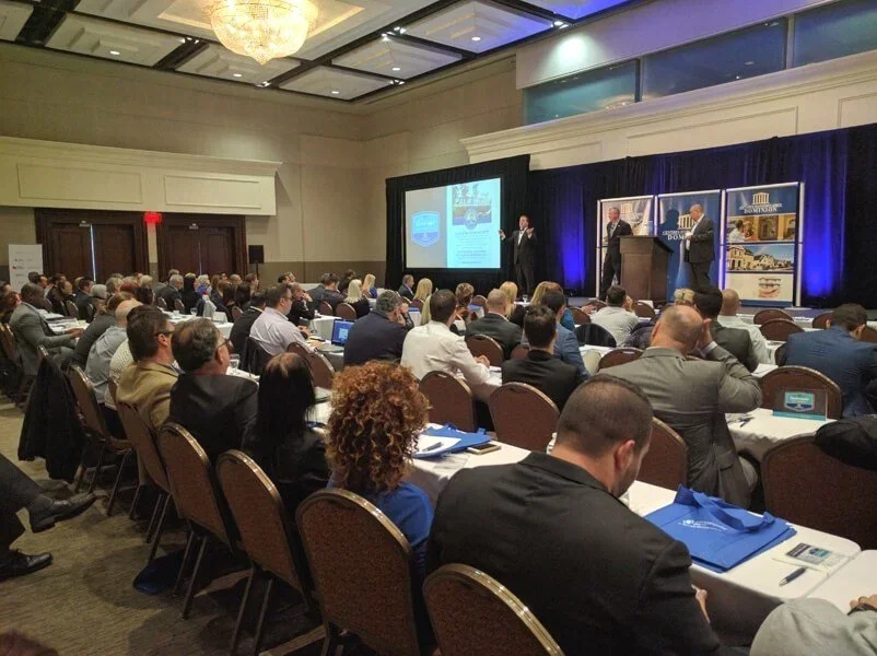 A large conference room filled with people sitting at tables, watching a presentation on stage with three speakers, a large screen, and banners.