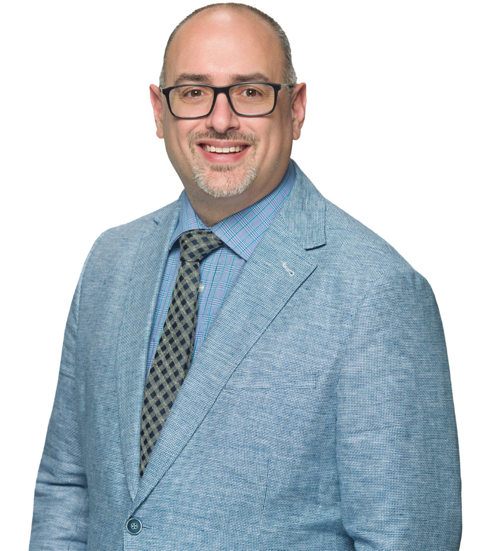 A professional man with glasses, a light blue blazer, a blue plaid shirt, and a patterned tie, smiling against a black background.
