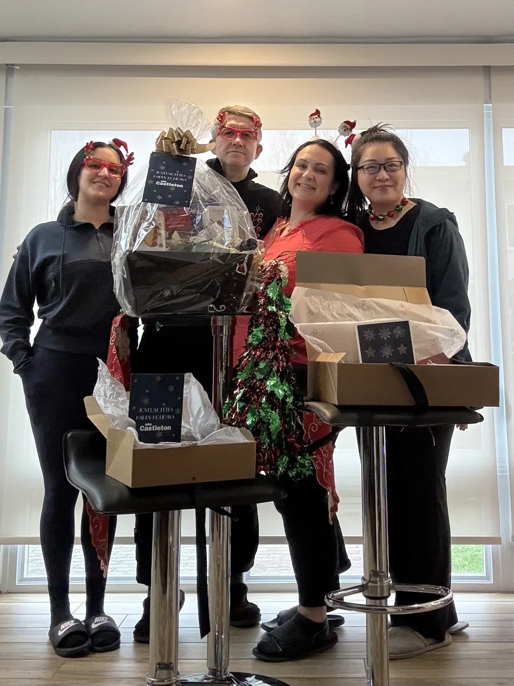 Four people celebrating Christmas indoors, standing behind two tables with gift boxes and wrapping paper, with Christmas decorations such as a small tinsel tree and festive headbands.