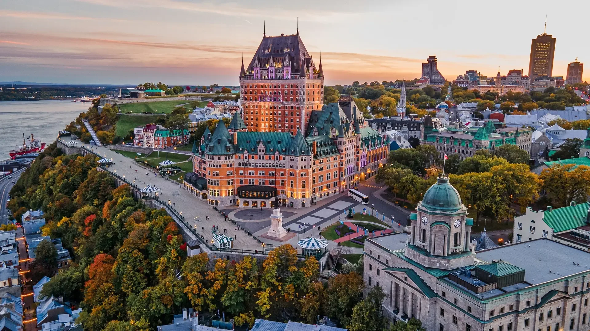 An aerial view of the Fairmont Le Château Frontenac hotel on a hill in Quebec City during sunset, with surrounding historic buildings, a clock tower, colorful autumn trees, and the St. Lawrence River in the background.