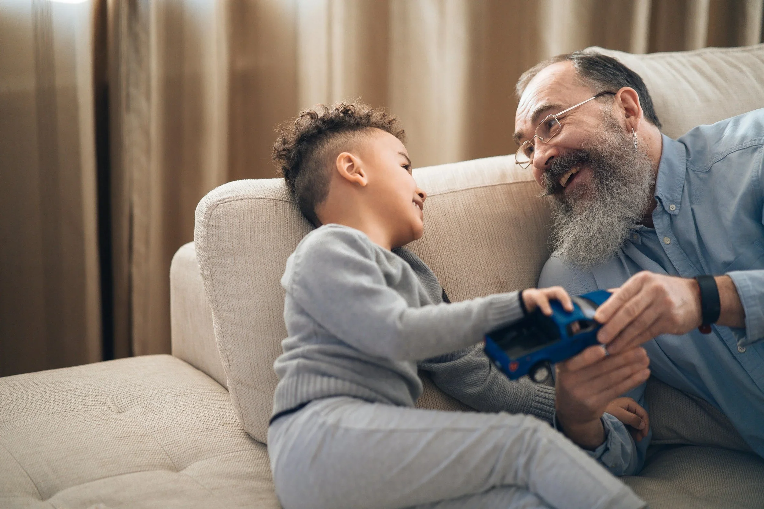 A young boy and an older man, likely his grandfather, are lying on a beige sofa, smiling and laughing while playing with a blue toy truck together.