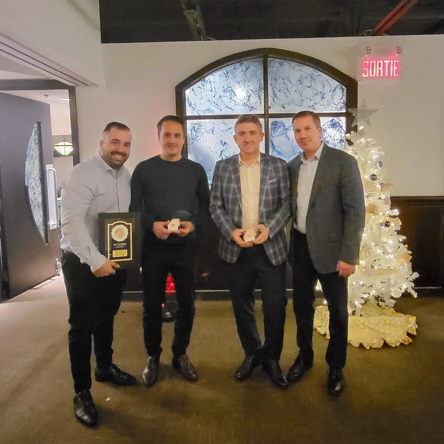 Four men standing in a row indoors, with a decorated Christmas tree and window behind them. The man on the far left is holding a plaque, the second man is holding a small gift box, the third man is holding a small object, and the fourth man is not ho