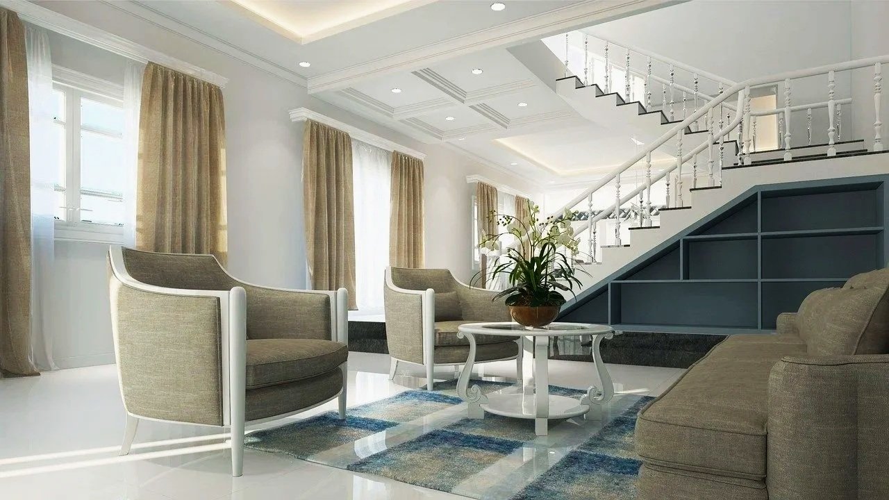Living room with beige armchairs and a sofa, a white coffee table with a potted plant, large windows with beige curtains, and a staircase with white railing and dark stairs.