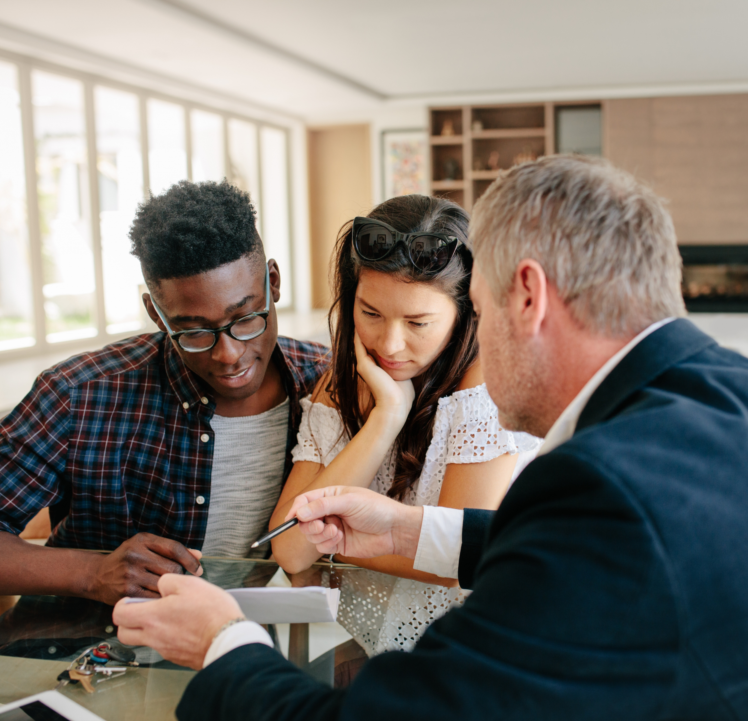 Three people, two young adults and an older man, sit at a table looking at a smartphone together in a bright room with large windows.