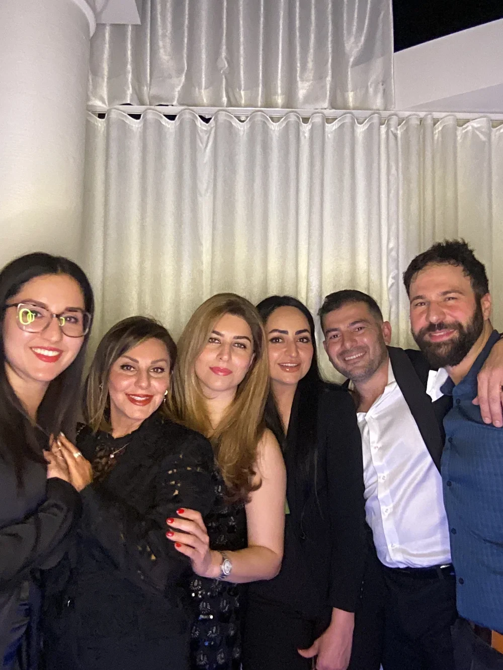 Six people standing together indoors, dressed in formal attire, smiling for a group photo against a backdrop of cream-colored curtains.