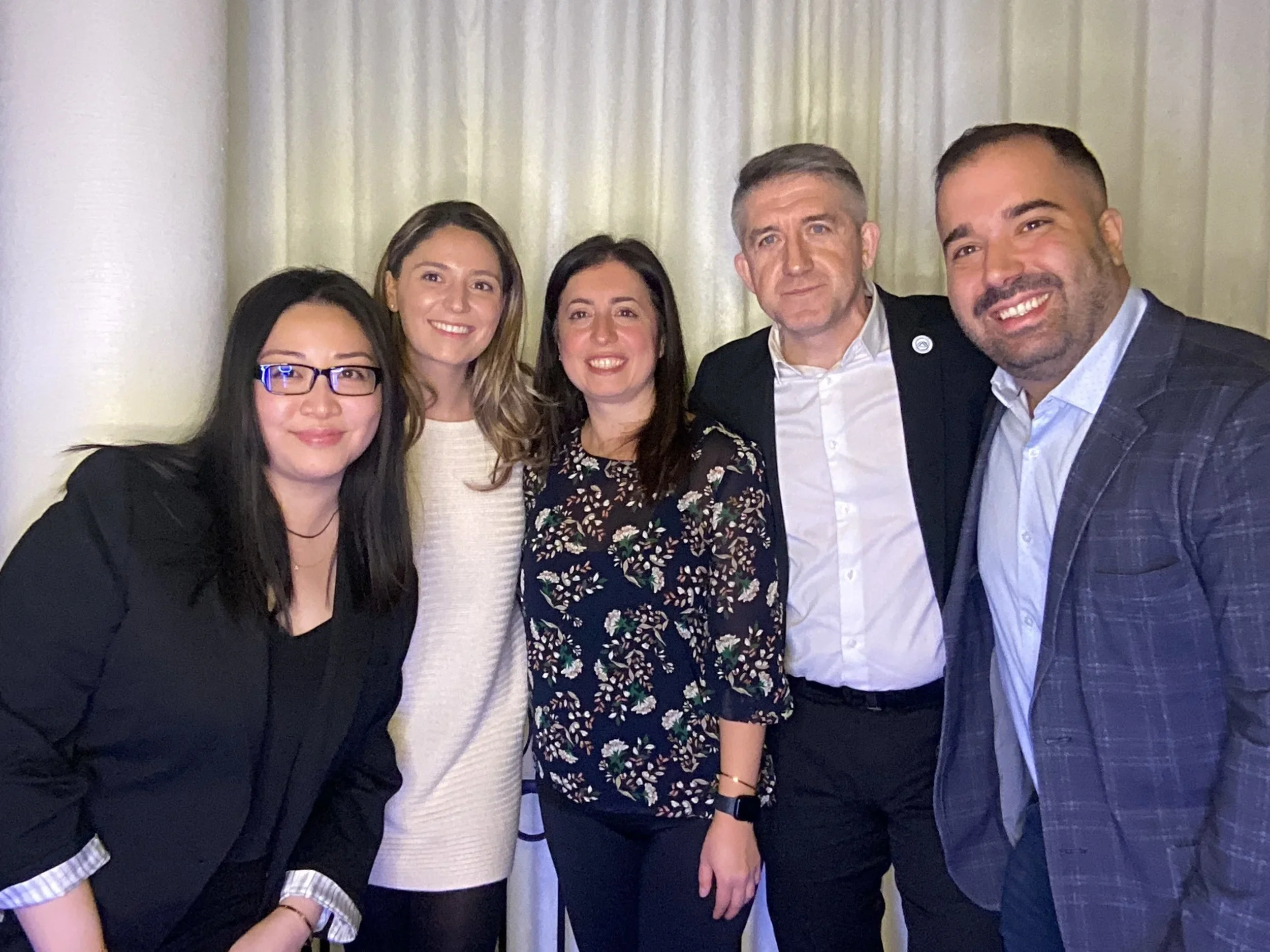 Group of five diverse people smiling and posing together indoors, dressed in business casual attire.