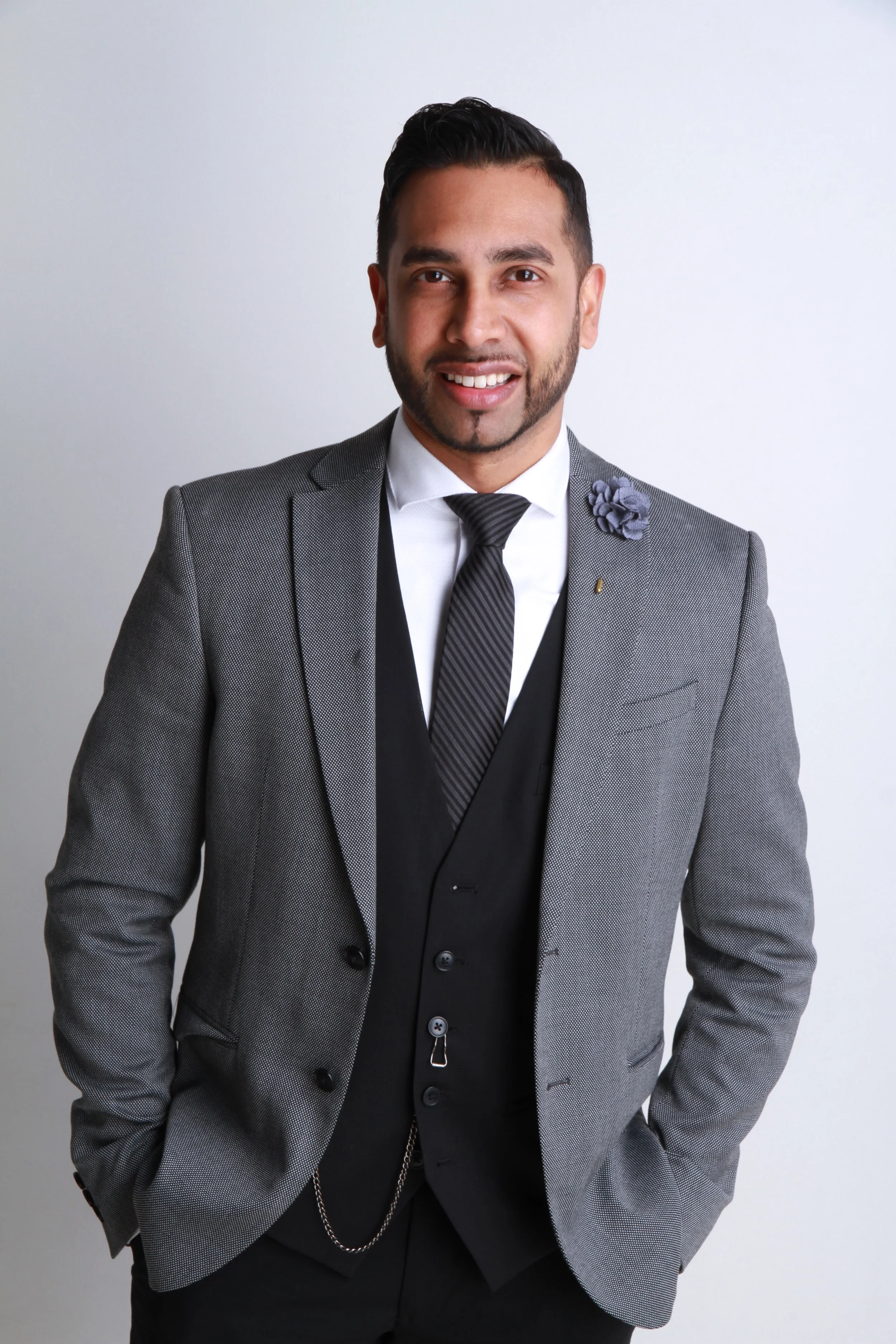 A man with a beard in a gray suit, white shirt, and striped tie, standing against a plain white background.