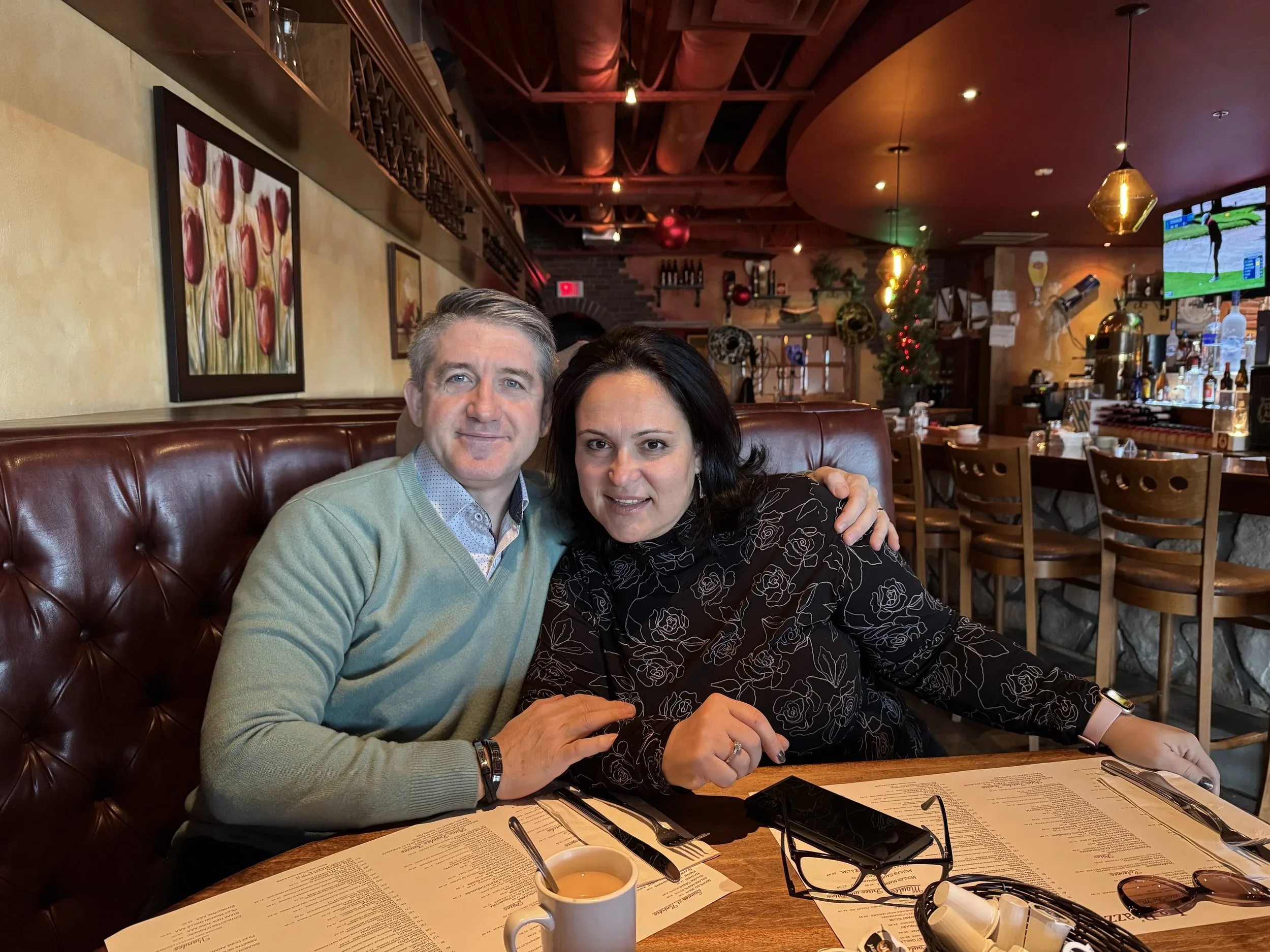 A man and a woman sitting together at a restaurant table, smiling at the camera. The table has a menu, a cup of coffee, glasses, and a smartphone. The background shows a decorated restaurant with paintings, a TV screen, and Christmas decorations.