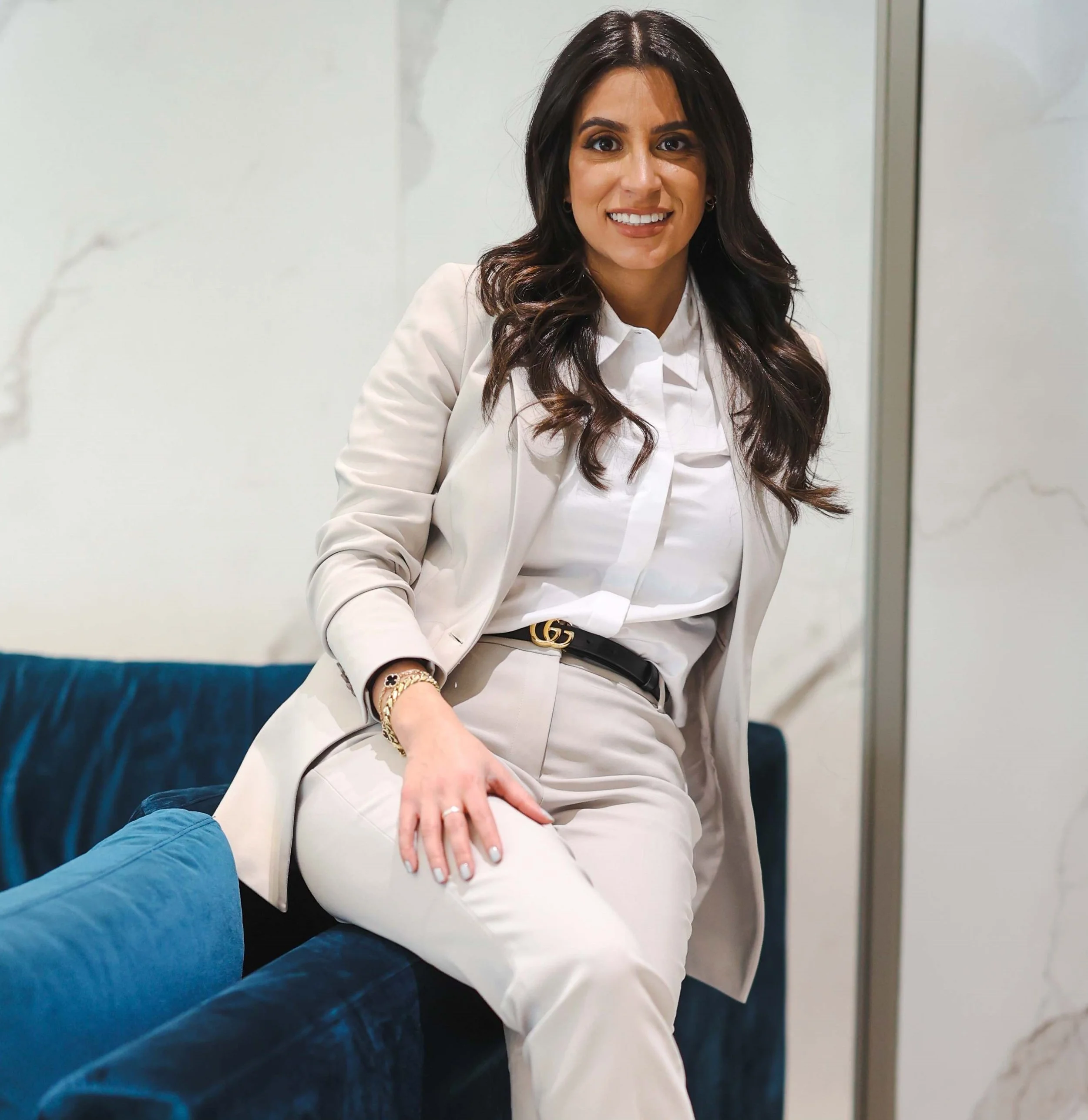 A woman with long dark hair wearing a white suit and white blouse, sitting on a blue velvet sofa, smiling, in a modern interior setting.