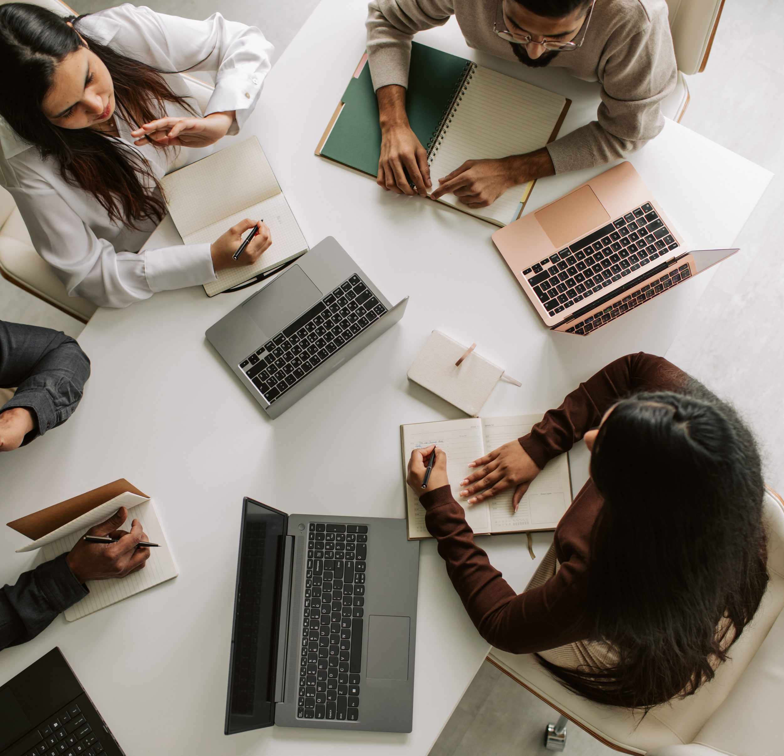 Top-down view of five people sitting around a white table with laptops, notebooks, and pens, engaged in a meeting or discussion.
