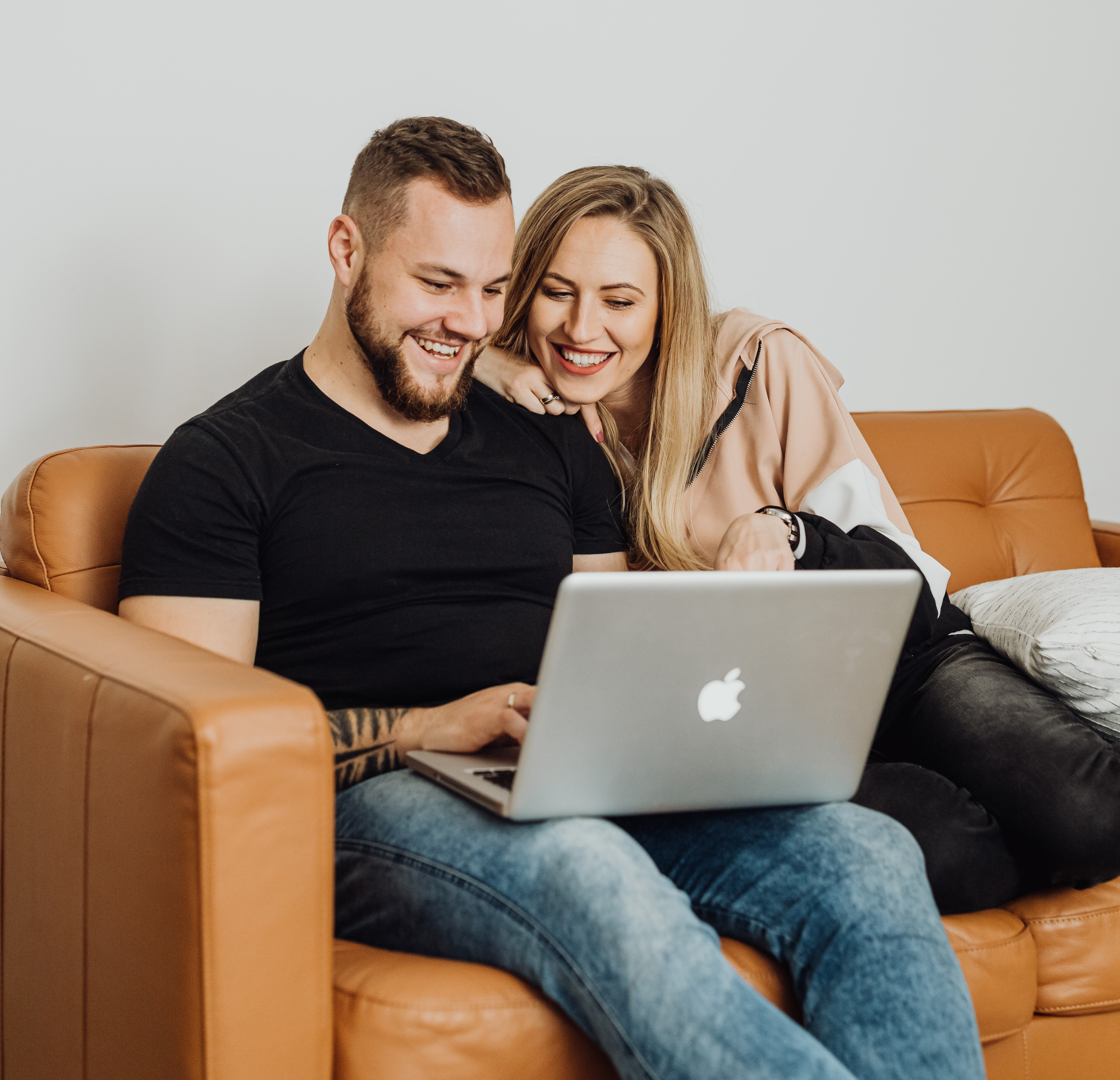 A young man and woman sitting on a tan leather couch, looking at a laptop and smiling, in a cozy living room.