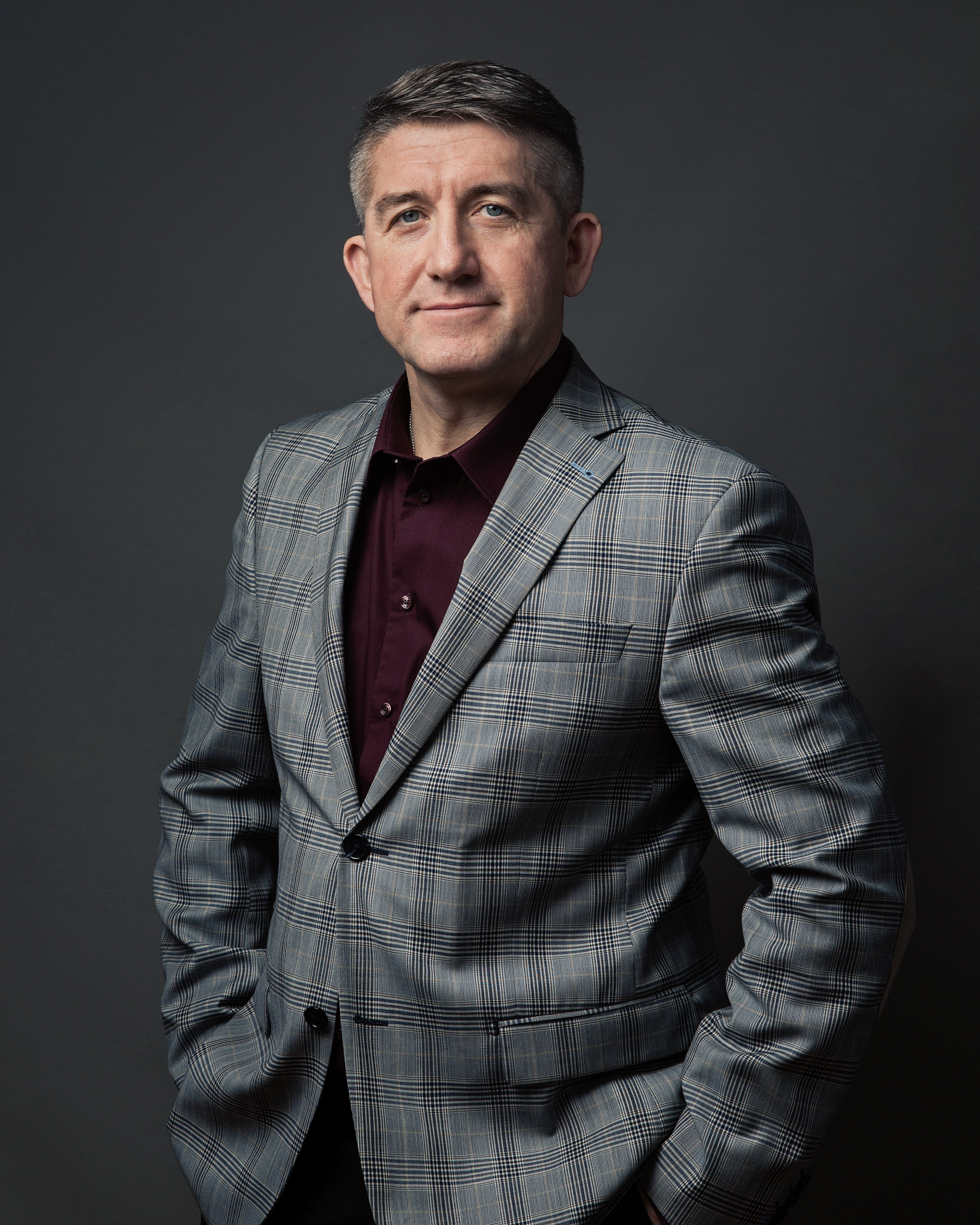 A middle-aged man with short gray hair wearing a gray plaid blazer over a maroon shirt, standing against a dark gray background.