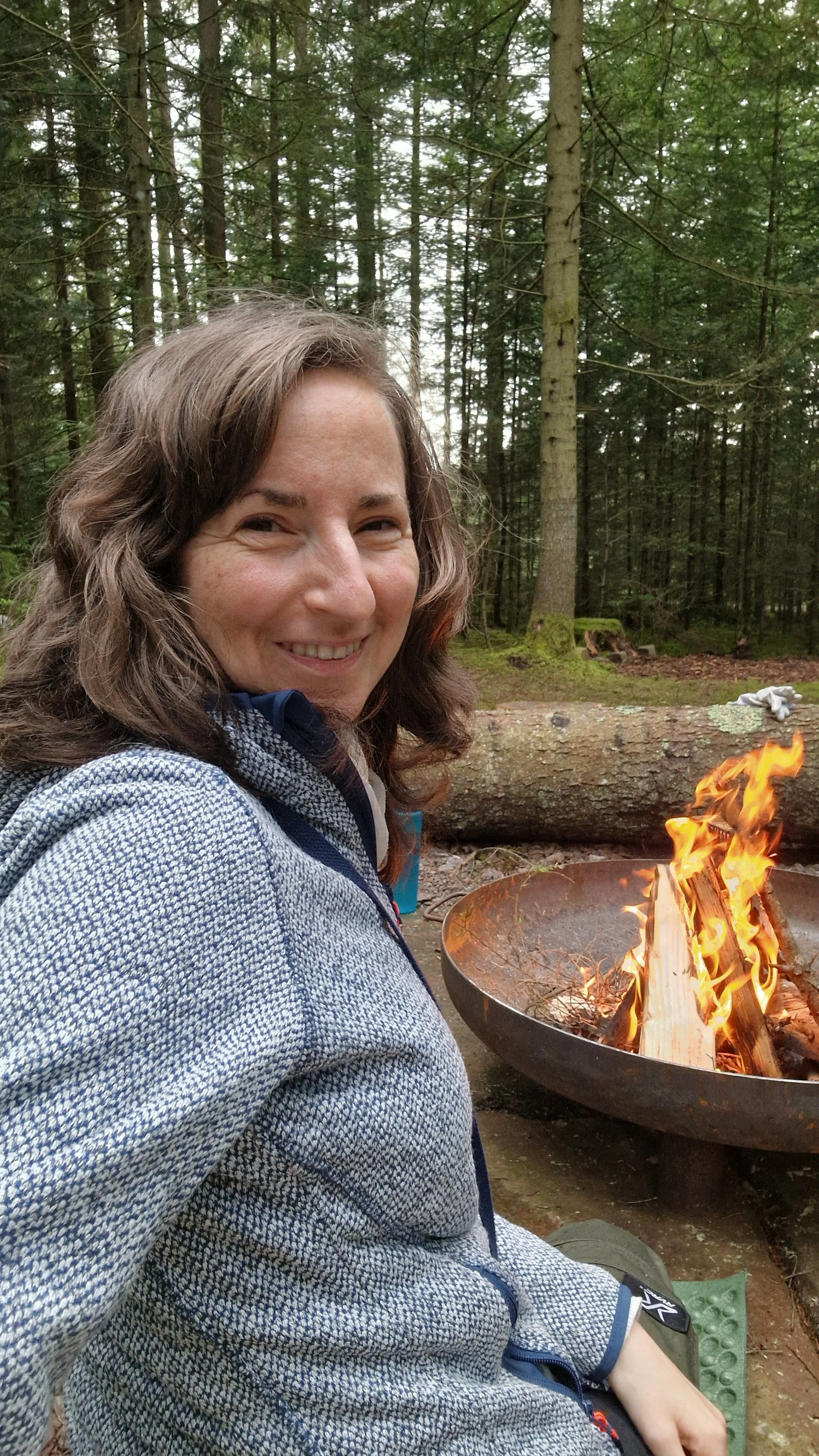 Eine lächelnde Frau sitzt neben einem Lagerfeuer in einem Wald, umgeben von Bäumen.