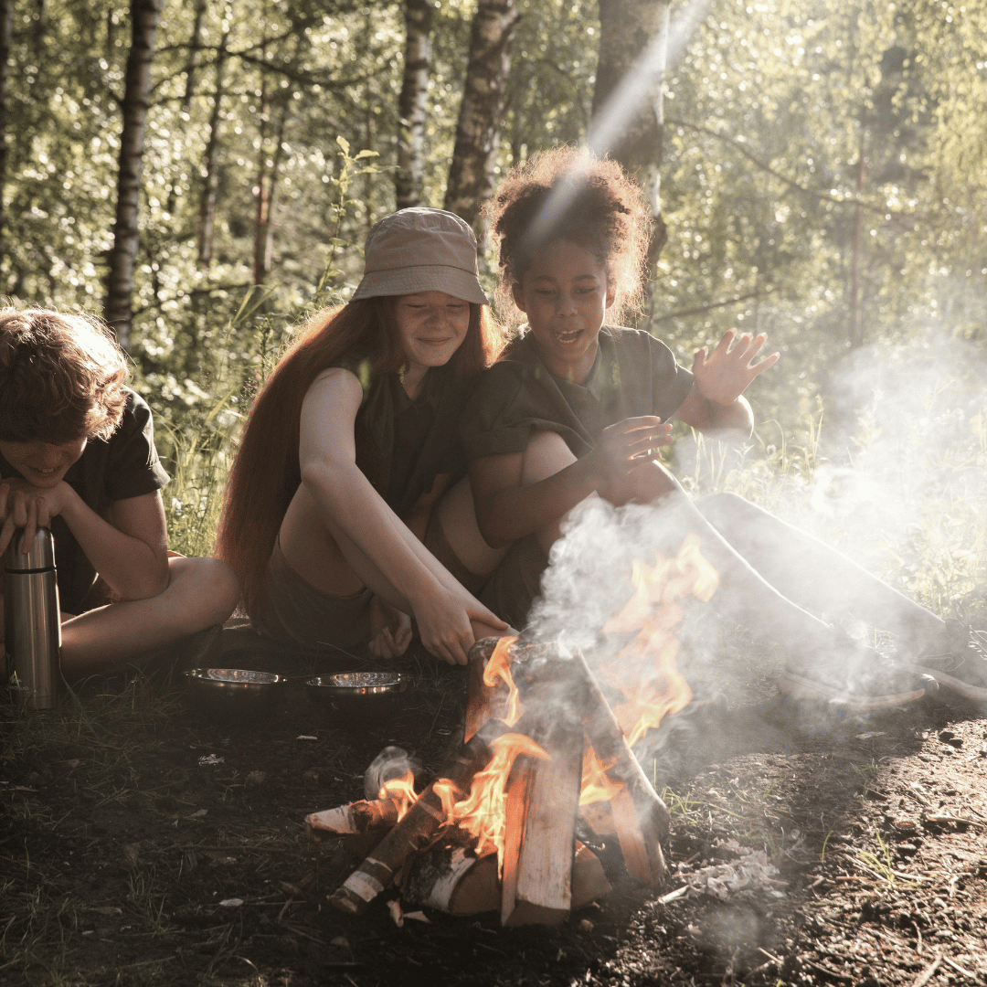 Drei Kinder sitzen am Lagerfeuer im Wald, beobachten die Flammen und lachen.