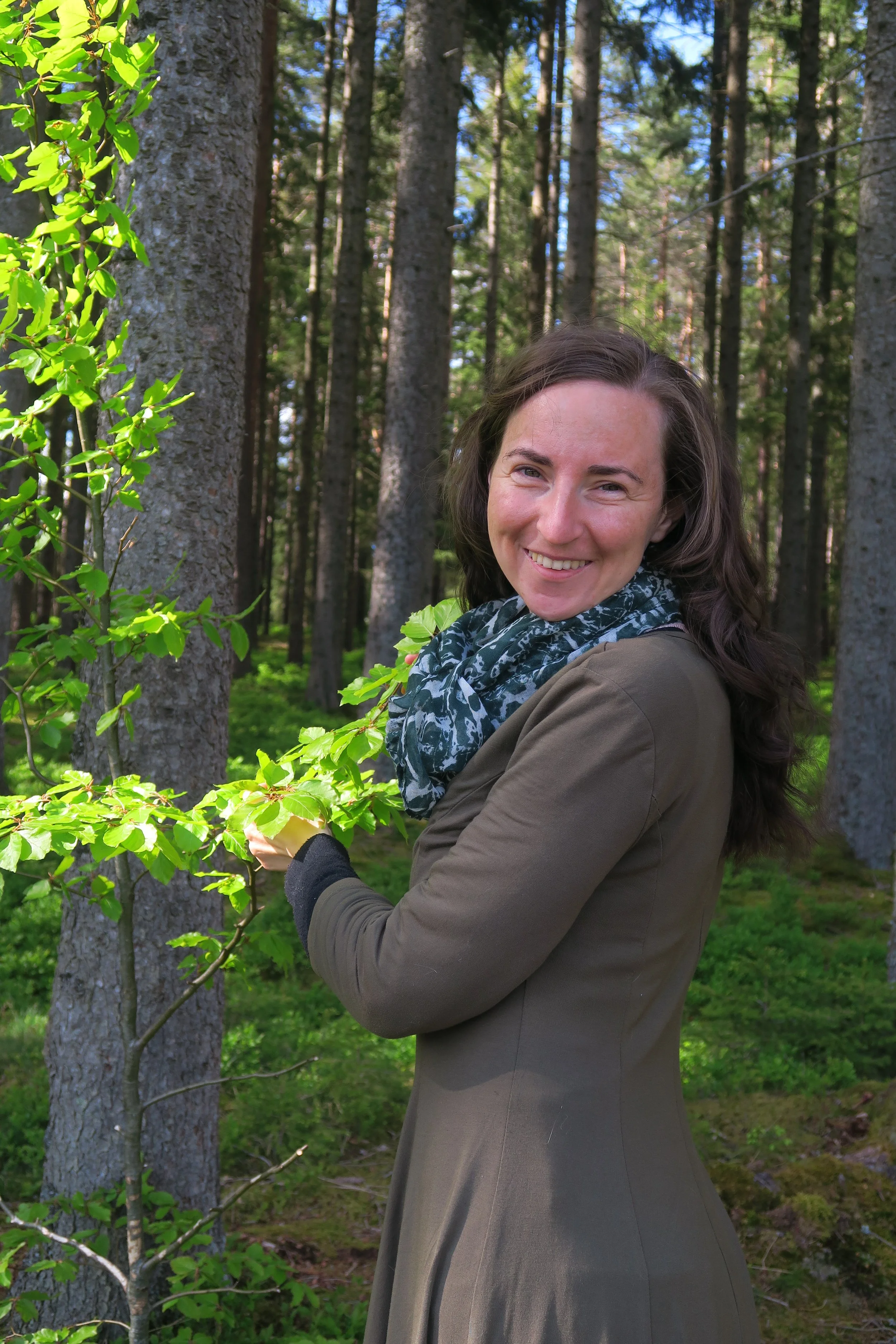 Eine lächelnde Frau in einem Wald, umgeben von Bäumen mit Unterholz.