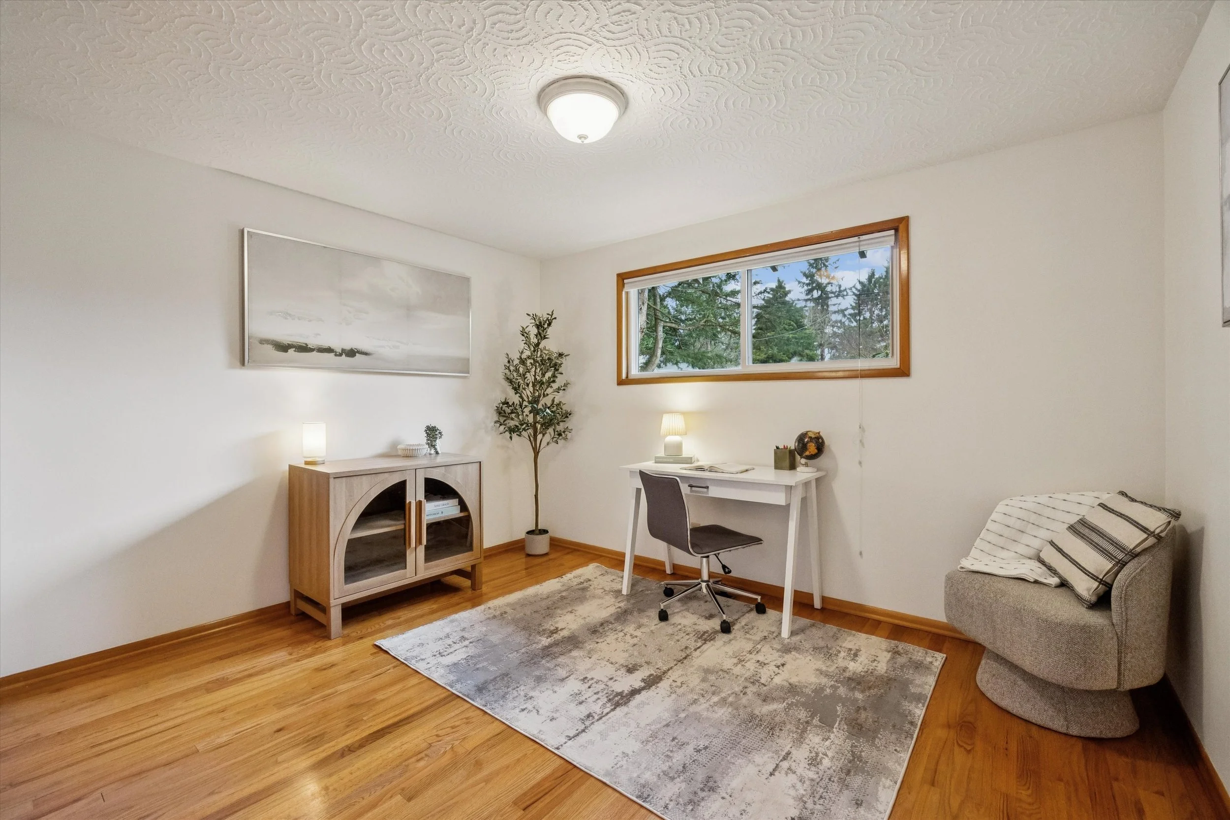 A minimalist home office with a white desk, gray office chair, beige armchair, wooden cabinet, potted plant, window view of trees, and abstract wall art.