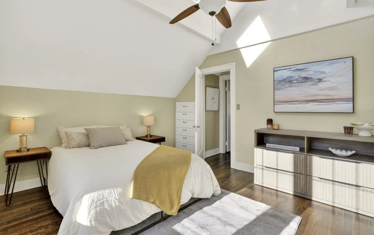 Bedroom with a sloped ceiling, bed with white bedding and gray pillows, wooden side tables with lamps, a dresser with decor, and a wall-mounted TV, in a room with hardwood flooring and a ceiling fan.