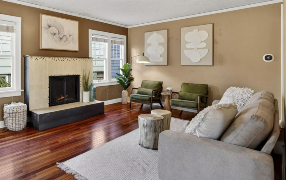 Living room with beige walls, hardwood floor, and a cozy seating area including a sofa and two green armchairs. A fireplace with a tiled surround is on the left, decorated with vases and a plant. Artwork hangs above the seating area.