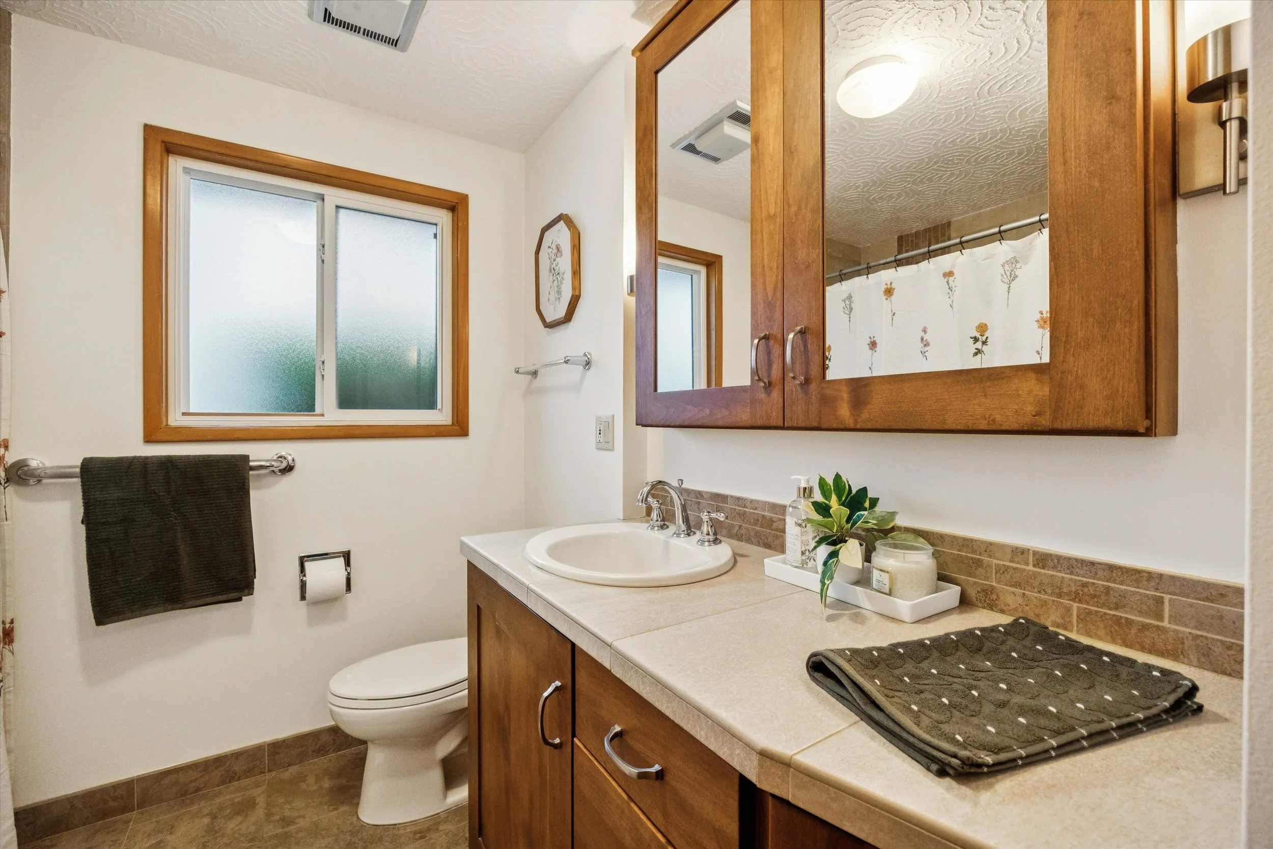 A bathroom with a window, a toilet, a wooden cabinet with mirror doors, and a countertop with a sink, soap, plants, and towels.