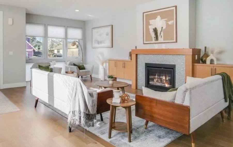 Living room with a gray sofa, white armchairs, a wooden coffee table, a brick fireplace, and wall art, with hardwood floors and a patterned area rug.
