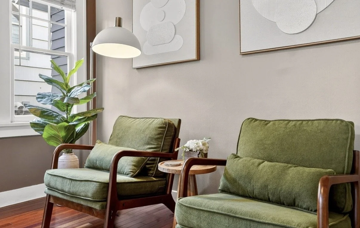 Living room corner with two green cushioned armchairs, a potted plant, a small wooden side table with flowers, a white hanging lamp, and abstract wall art, next to a window with exterior siding visible.