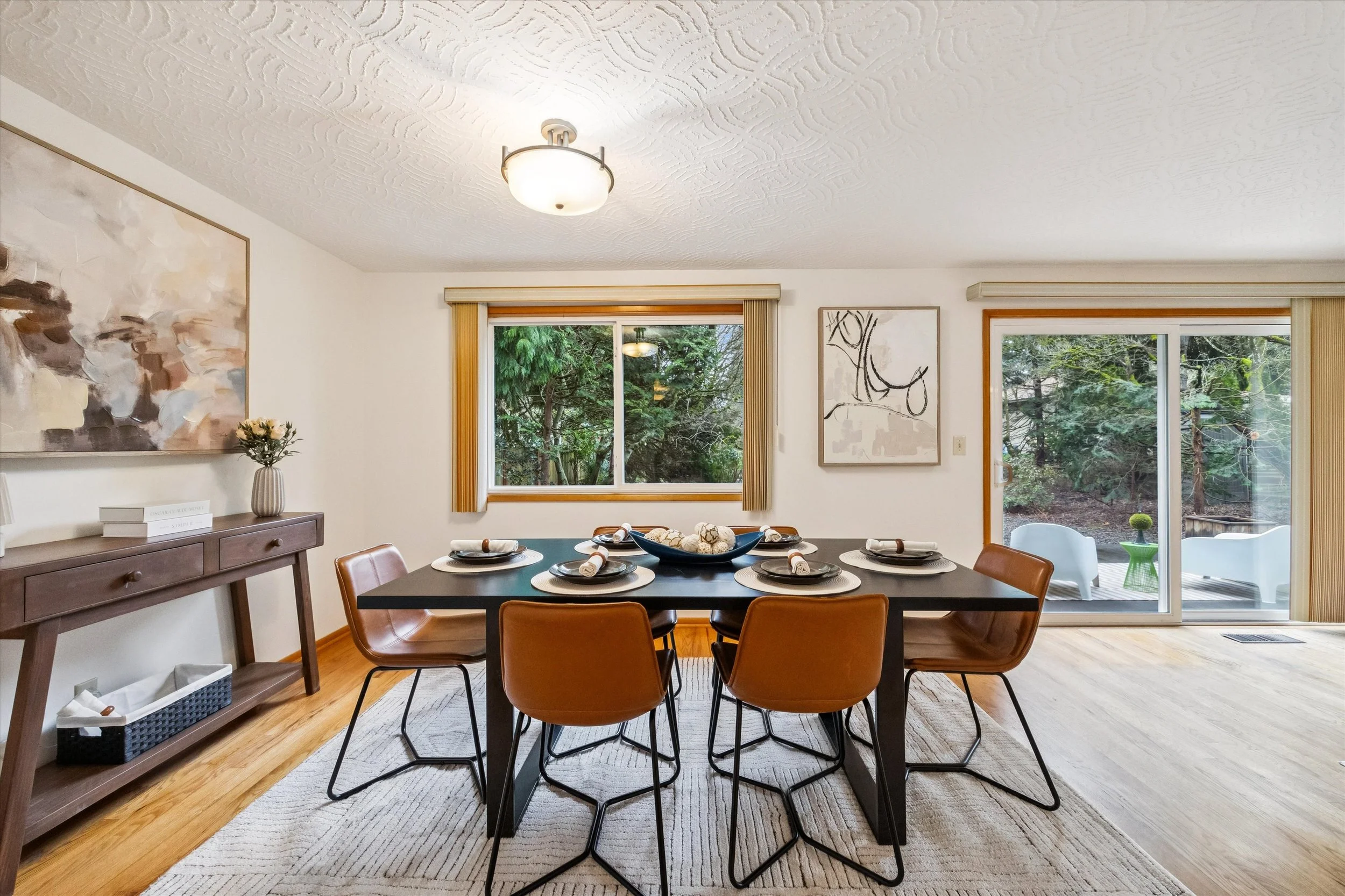 Dining room with a black table set for six, brown chairs, a sideboard, abstract artworks, large window with views of greenery, and a sliding glass door to an outdoor patio.