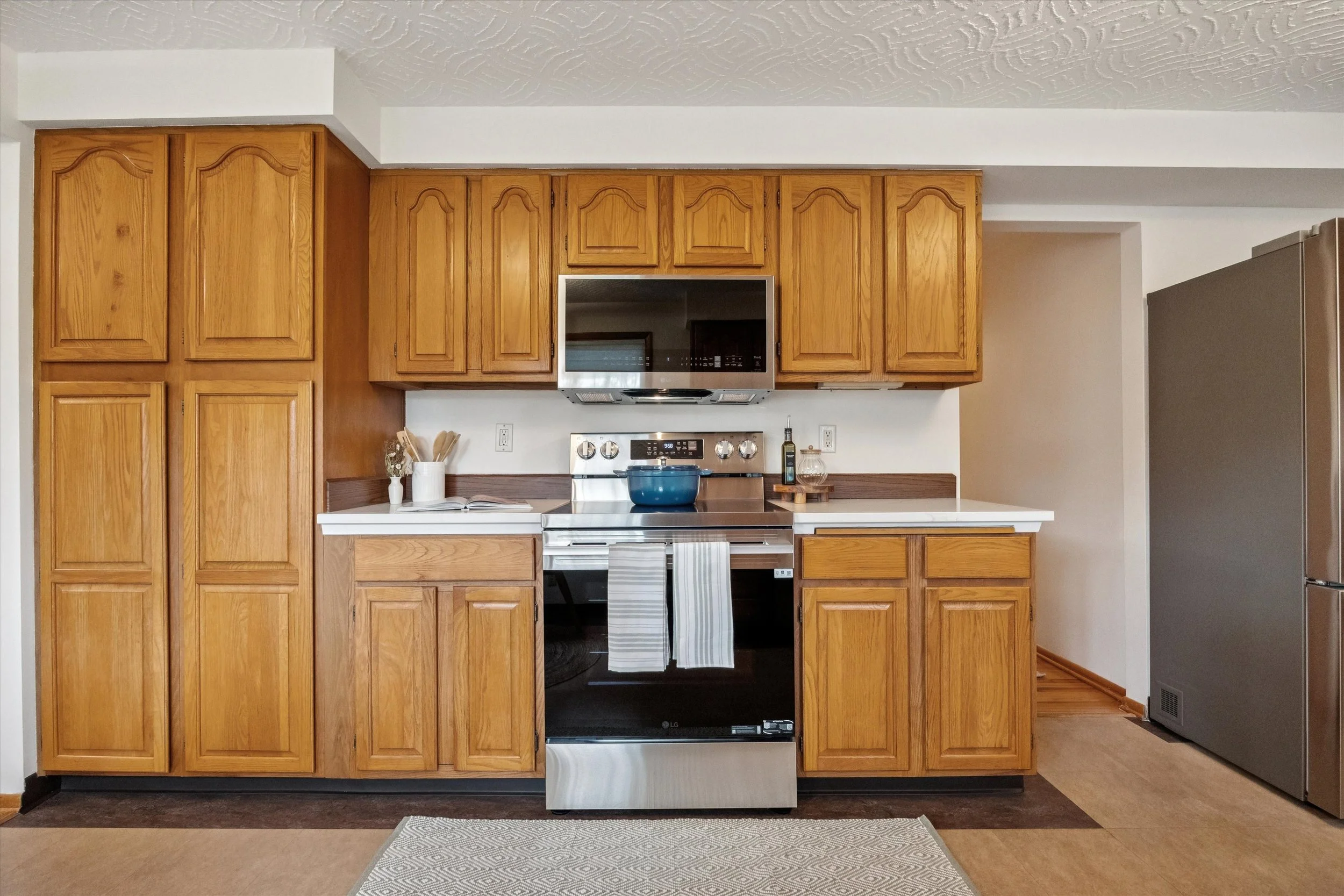 Kitchen with wooden cabinets, stainless steel stove and microwave, black dishwasher, and a large silver refrigerator. Countertop decor includes a blue pot, oil bottle, and small tray with bottles.