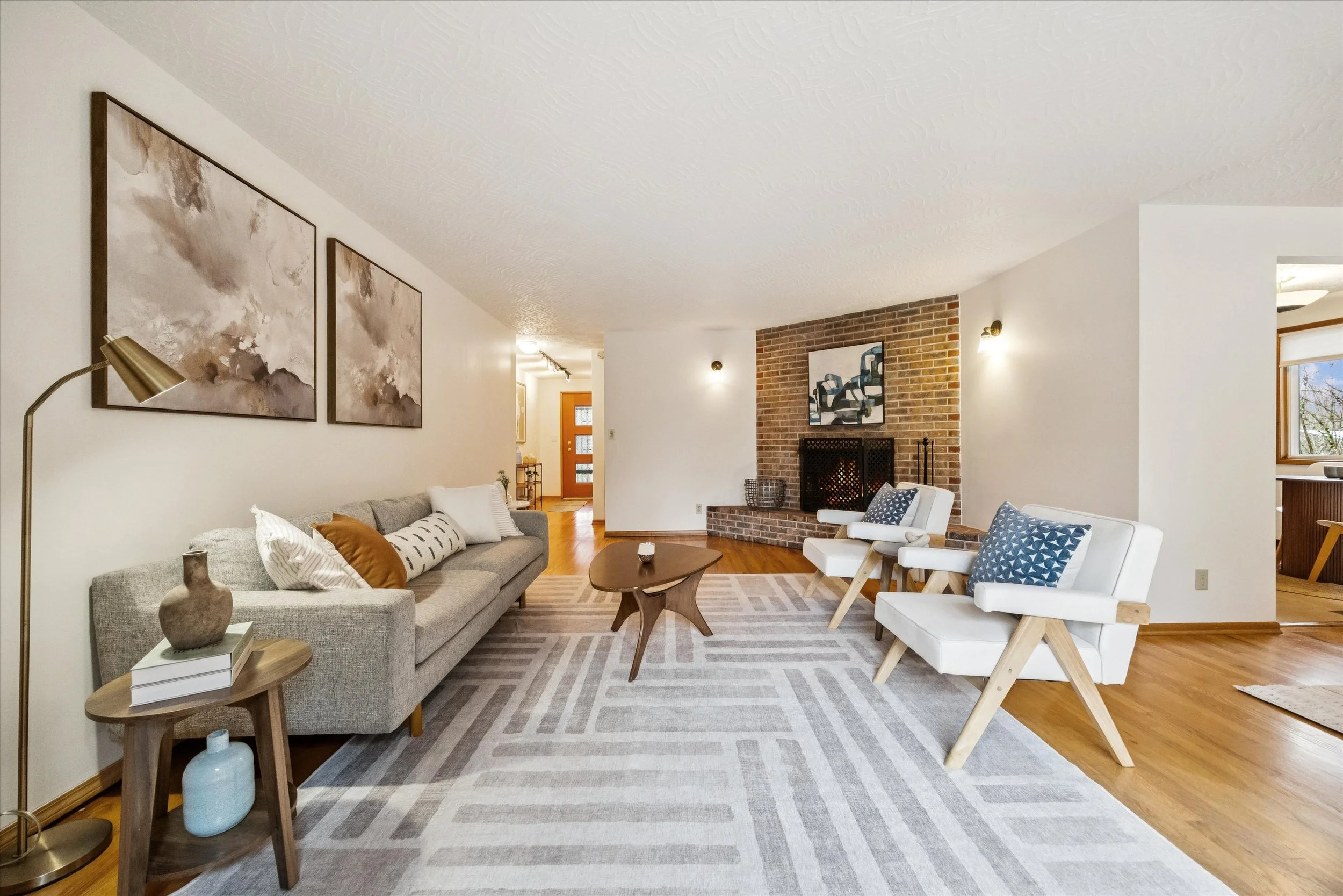 Living room with a beige sofa, two white armchairs, a wooden coffee table, a fireplace, and wall art, with hardwood floors and a light gray patterned rug.