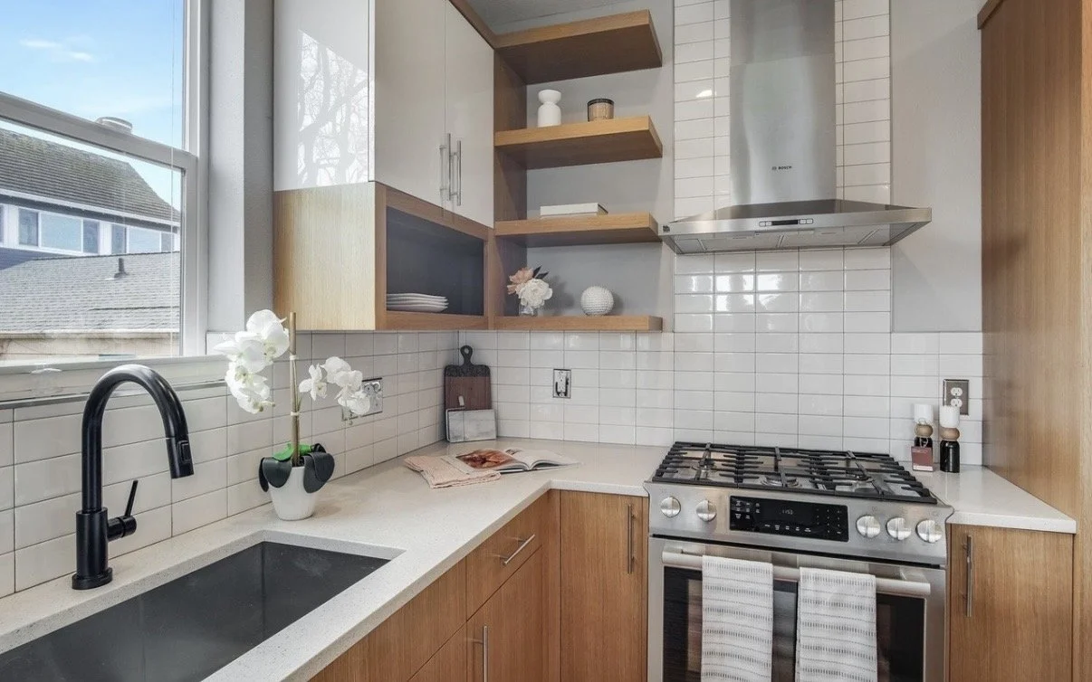 A modern kitchen with wooden cabinets, open shelves, a black sink, a gas stove, and a window with a view of neighboring houses and trees.