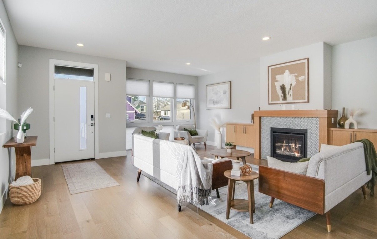 Living room with a white couch, armchair, and coffee table, fireplace with artwork above, large windows, wooden flooring, and decor including vases and plants.