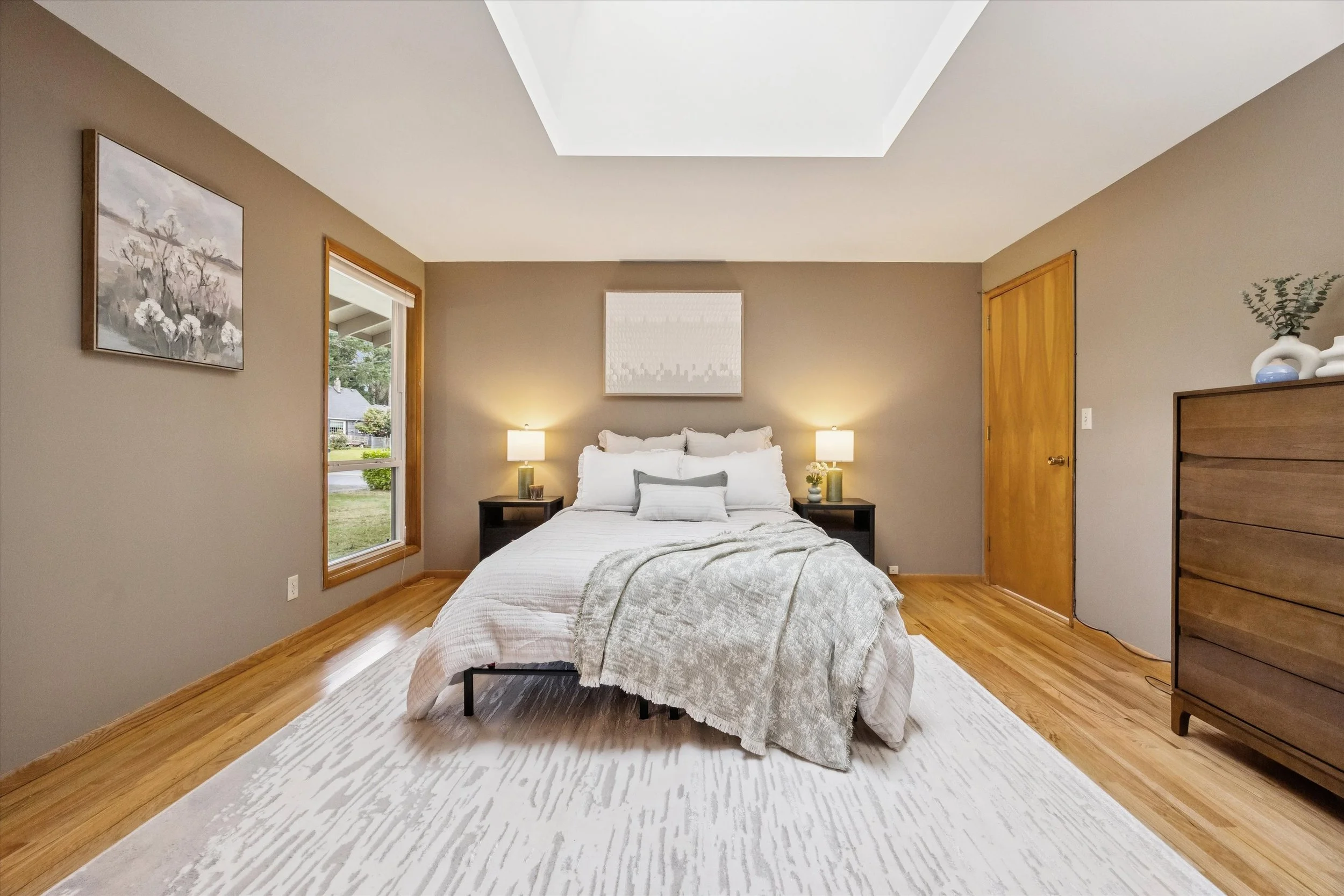 A cozy bedroom with a bed, two nightstands with lamps, a window, a painting above the bed, and a wooden dresser on the right, with hardwood flooring and a white rug.
