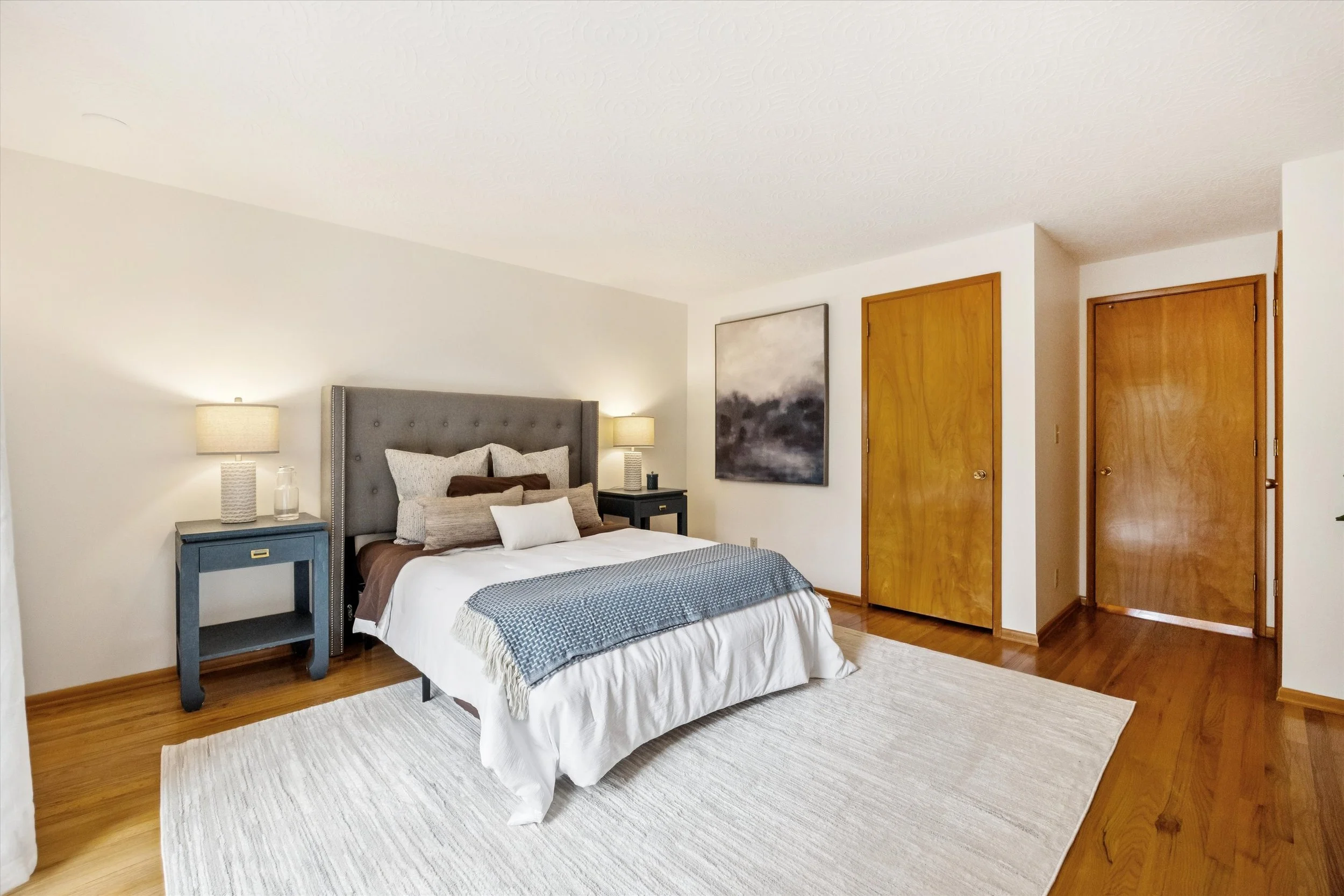 A bedroom with a bed, two nightstands with lamps, a painting on the wall, and wooden doorways.