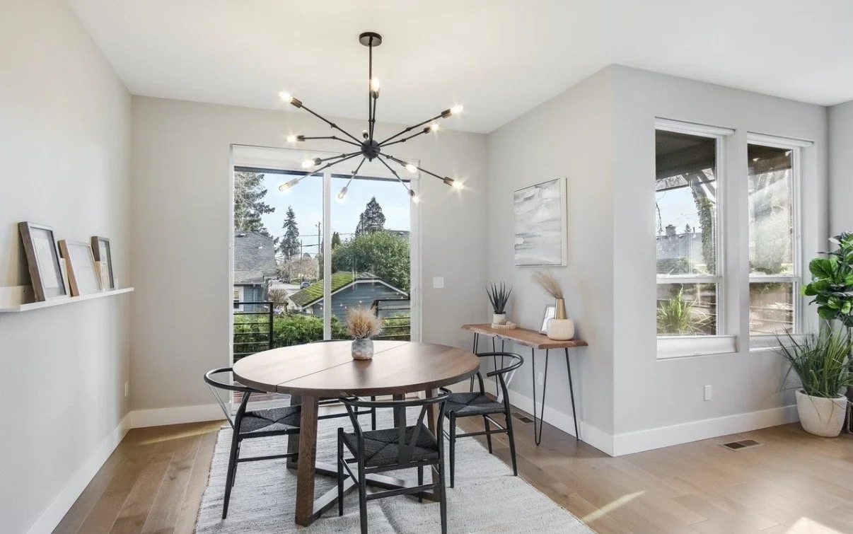 Bright dining room with a round wooden table, surrounded by four black chairs, a modern chandelier overhead, and large windows letting in natural light. Decor includes framed art, a side table with plants, and a large potted plant in the corner.
