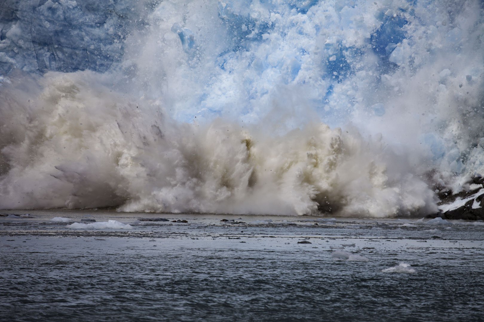 Ice breaking from Northwestern Glacier in Alaska, with a cloud of spray and debris rising above the water.