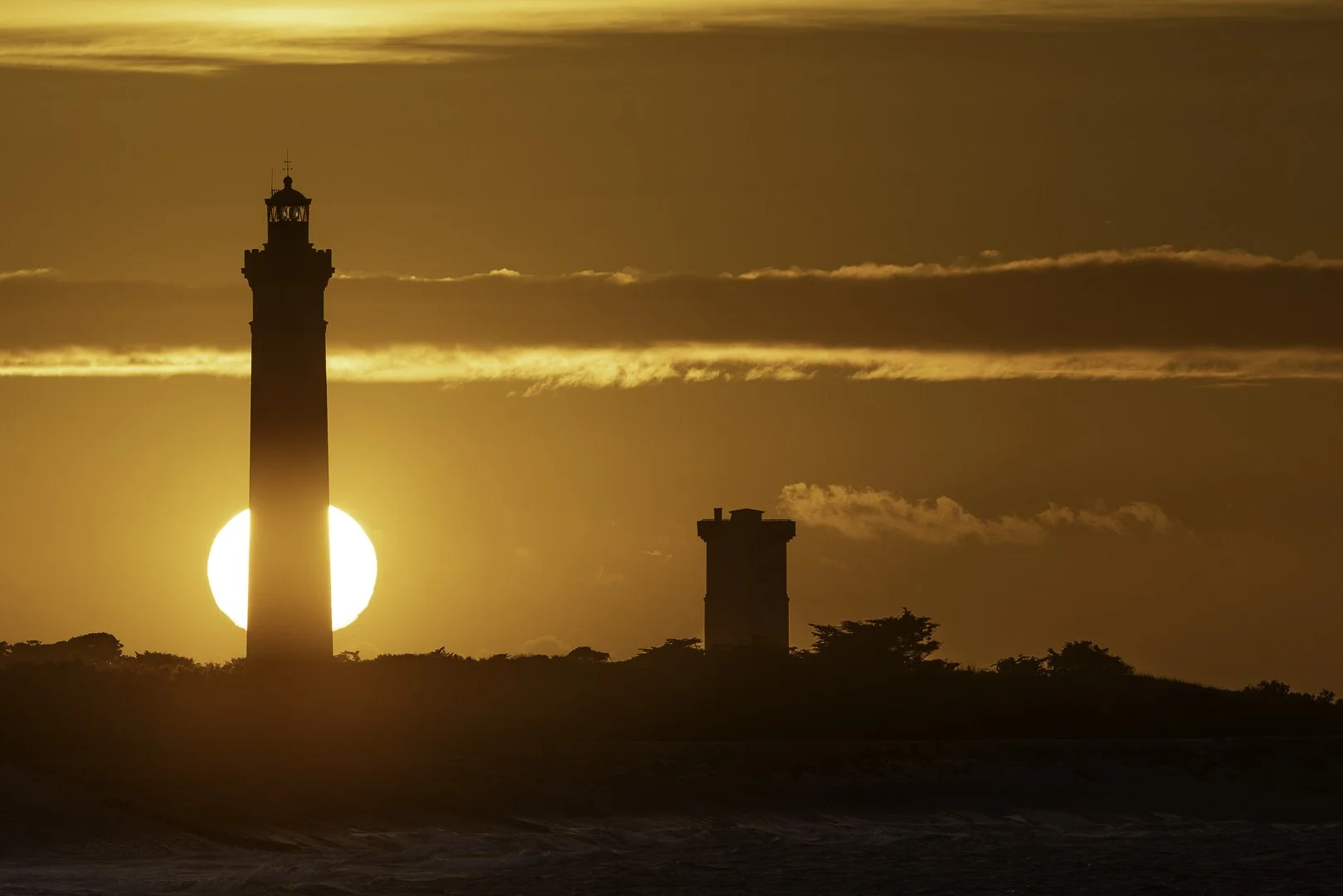 Sun setting directly behind the Phare des Baleines lighthouse on Île de Ré, France, creating a dramatic golden silhouette.
