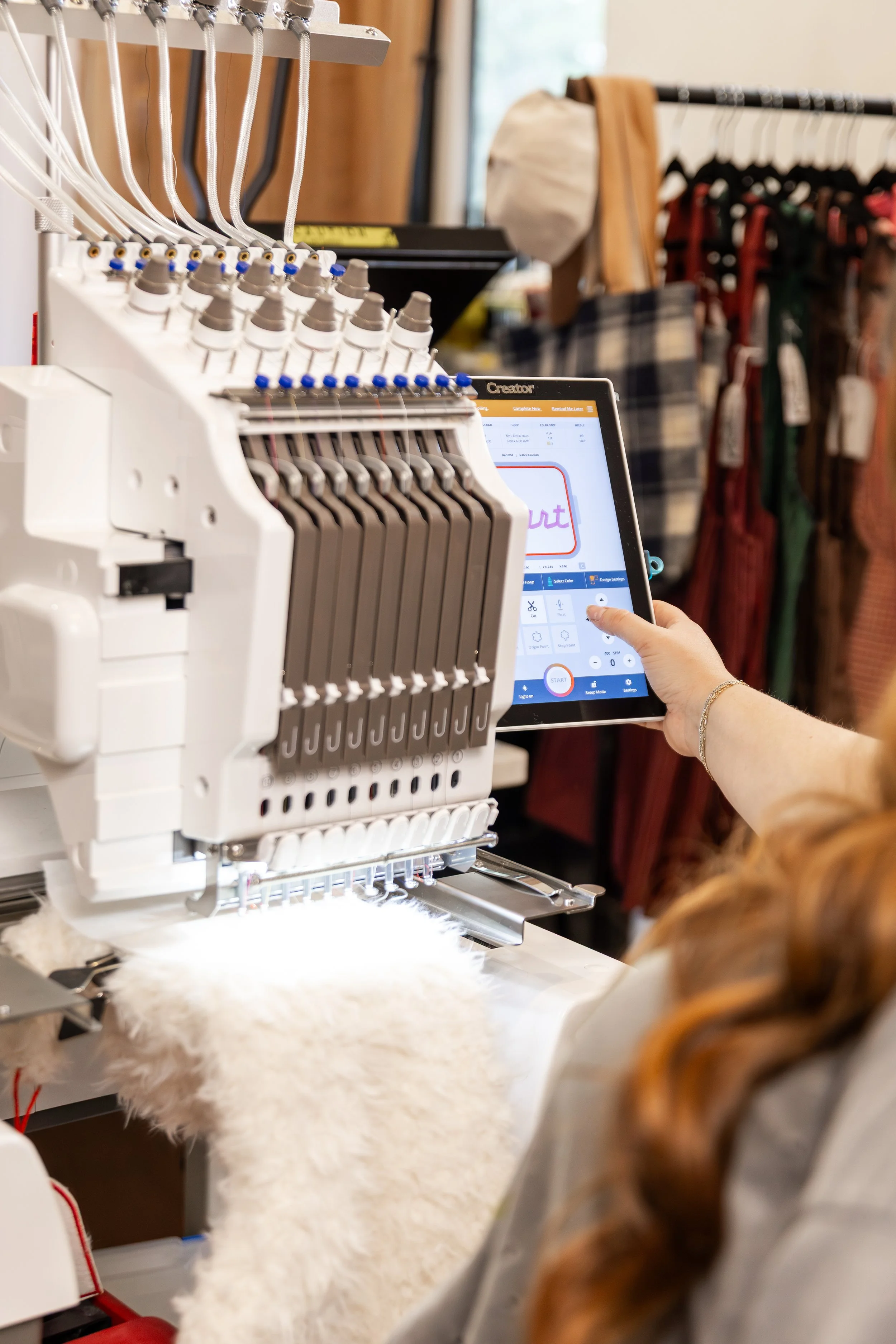A person using embroidery machine with digital touchscreen in a craft store, surrounded by clothing racks.