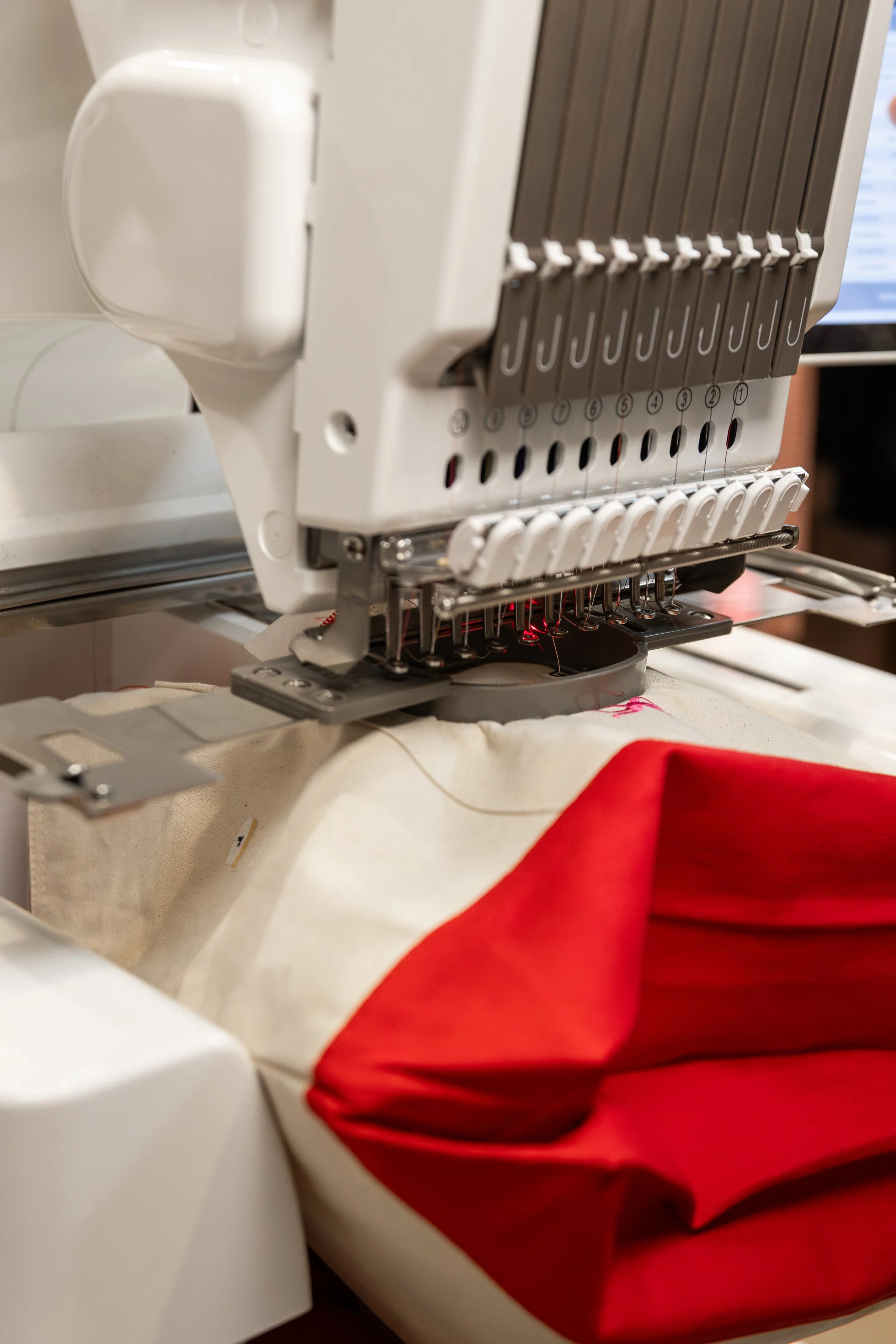 Embroidery machine stitching a red and white fabric with a computer monitor in the background.
