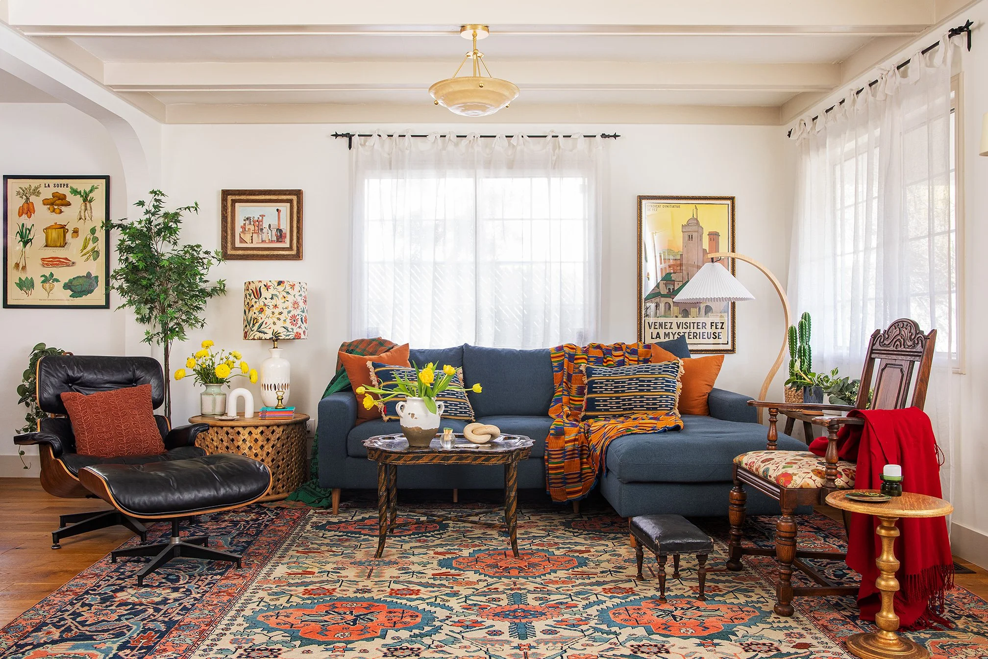 Living room with a blue sectional sofa, a black leather lounge chair, wooden side tables, patterned rugs, framed artwork, vintage decor, and white curtains.
