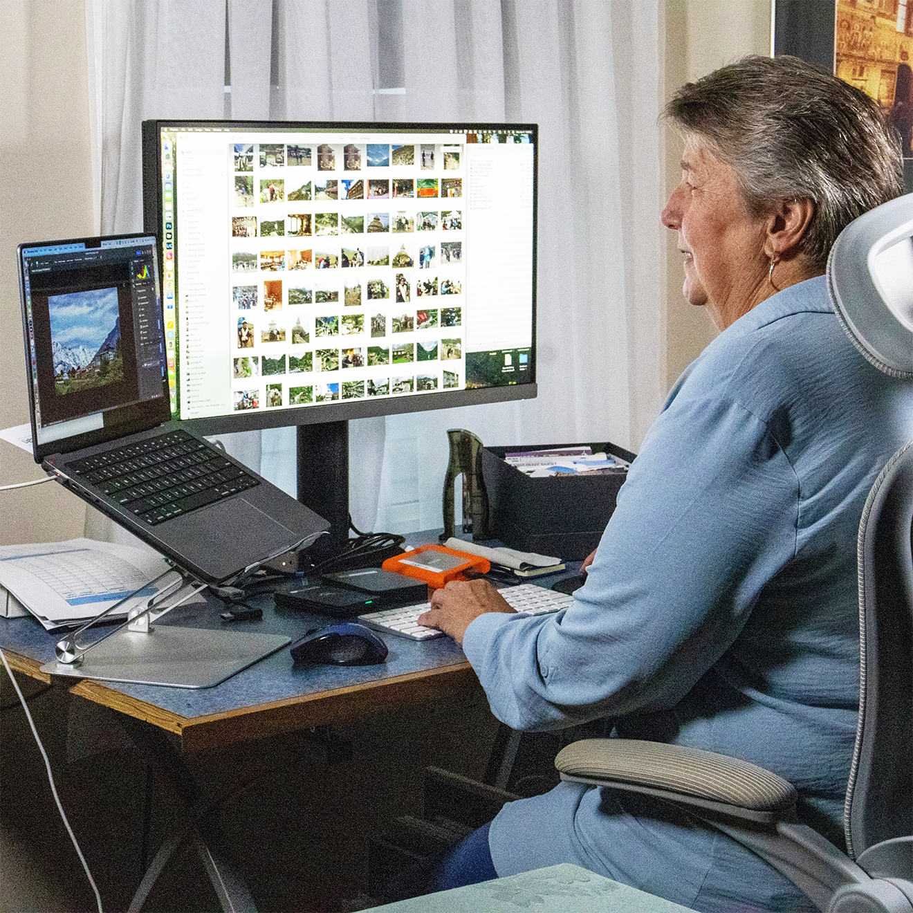 A woman sitting at a desk, looking at a computer monitor filled with photo thumbnails, with a laptop open to her left showing a landscape picture. The desk has various items including papers, a mouse, and an external drive.
