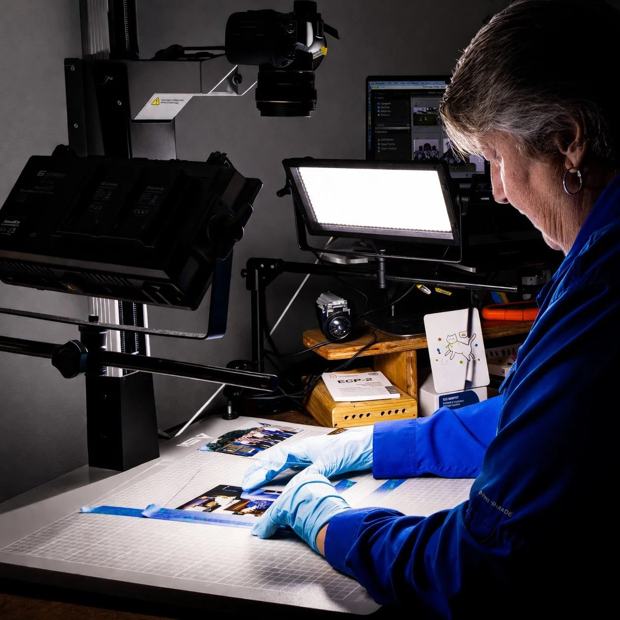 A person wearing gloves working on photographs or prints in a professional studio or lab, with lighting and camera equipment surrounding them.