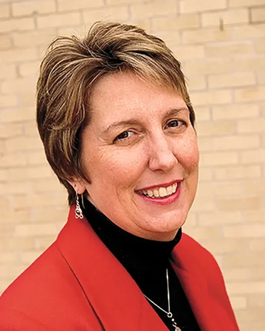 A woman with short, light brown hair smiling, wearing a red blazer and black top, against a light-colored brick wall background.