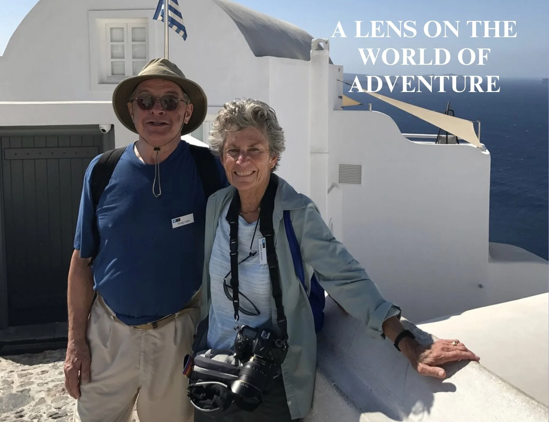 Two smiling older adults standing on a white stone balcony overlooking the blue ocean with white buildings and a Greek flag. The man wears a wide-brimmed hat, sunglasses, a blue shirt, and khaki pants. The woman wears a light jacket, has a camera han