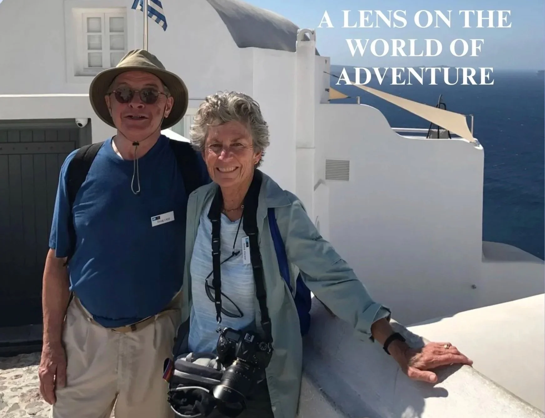 Two smiling tourists, a man with a wide-brimmed hat and sunglasses, and a woman with a camera around her neck, stand outdoors on a sunny day with a white building and blue sea in the background. Text on the image reads, 'A LENS ON THE WORLD OF ADVENTURE.'