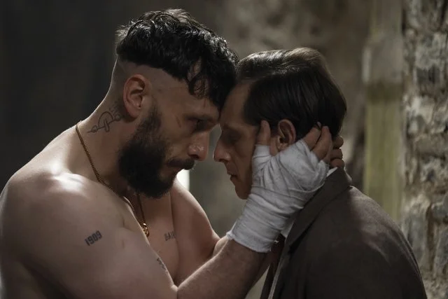 Reuben (Richard Gadd) holds Niall's (Jamie Bell's) face in his hands, which are taped for boxing. Richard Gadd is shirtless with multiple tattoos, and Niall is in traditional Scots formalwear.