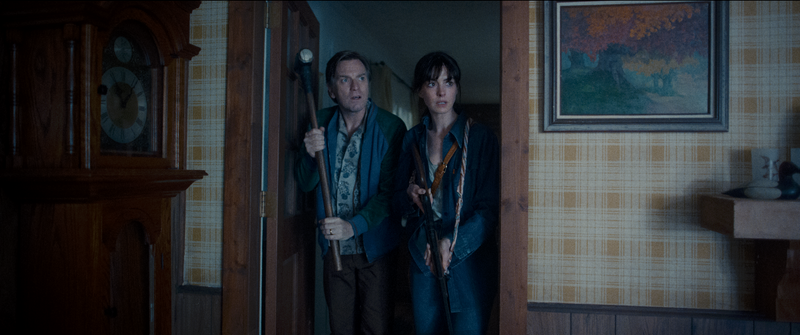 In David Robert Mitchell's 2026 film THE END OF OAK STREET, Greg (Ewan McGregor) and Denise (Anne Hathaway) enter their 1980s suburban living room doorway beside a grandfather clock, holding a sledgehammer and a rifle to defend themselves.