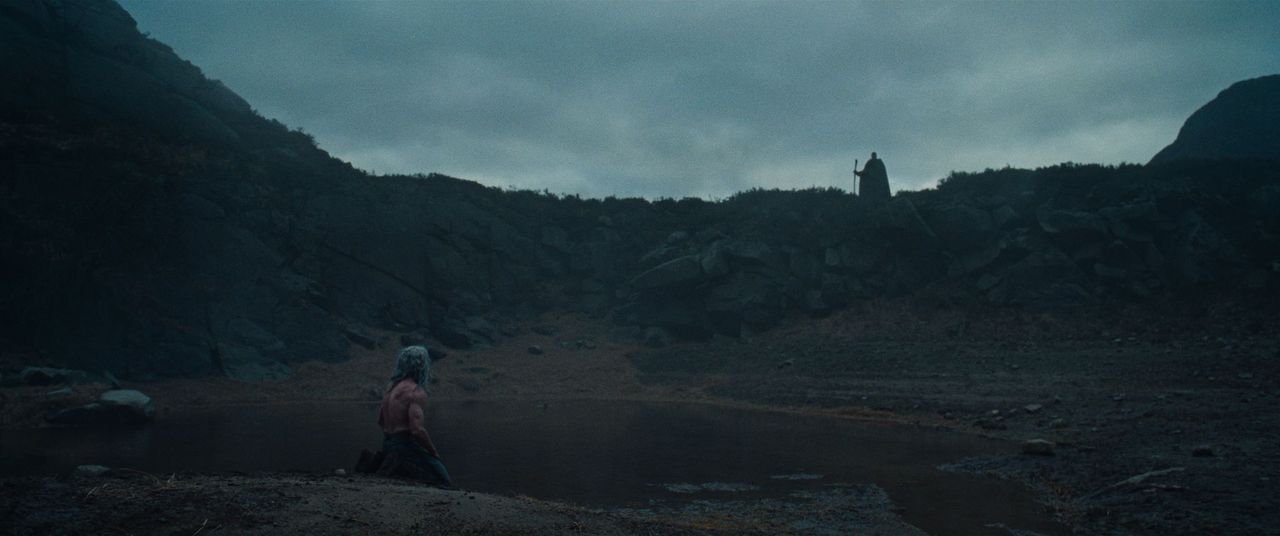 Robin Hood (Hugh Jackman) kneels shirtless at a body of water as a man on a distant cliff with a staff looks over him in THE DEATH OF ROBIN HOOD.